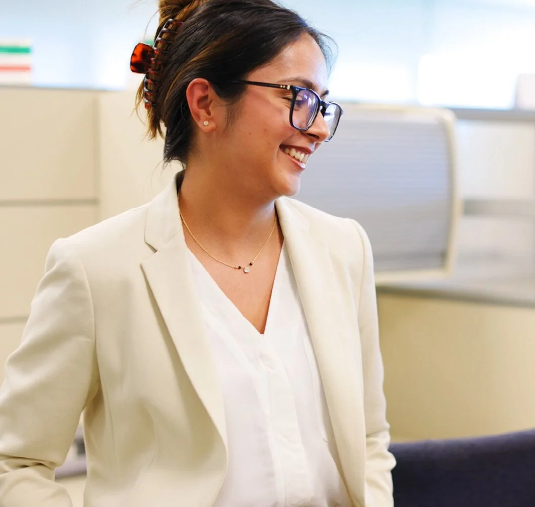A woman with short dark hair, glasses, and a hair clip, smiling and wearing a cream-colored blazer over a white blouse, sitting in an office setting.
