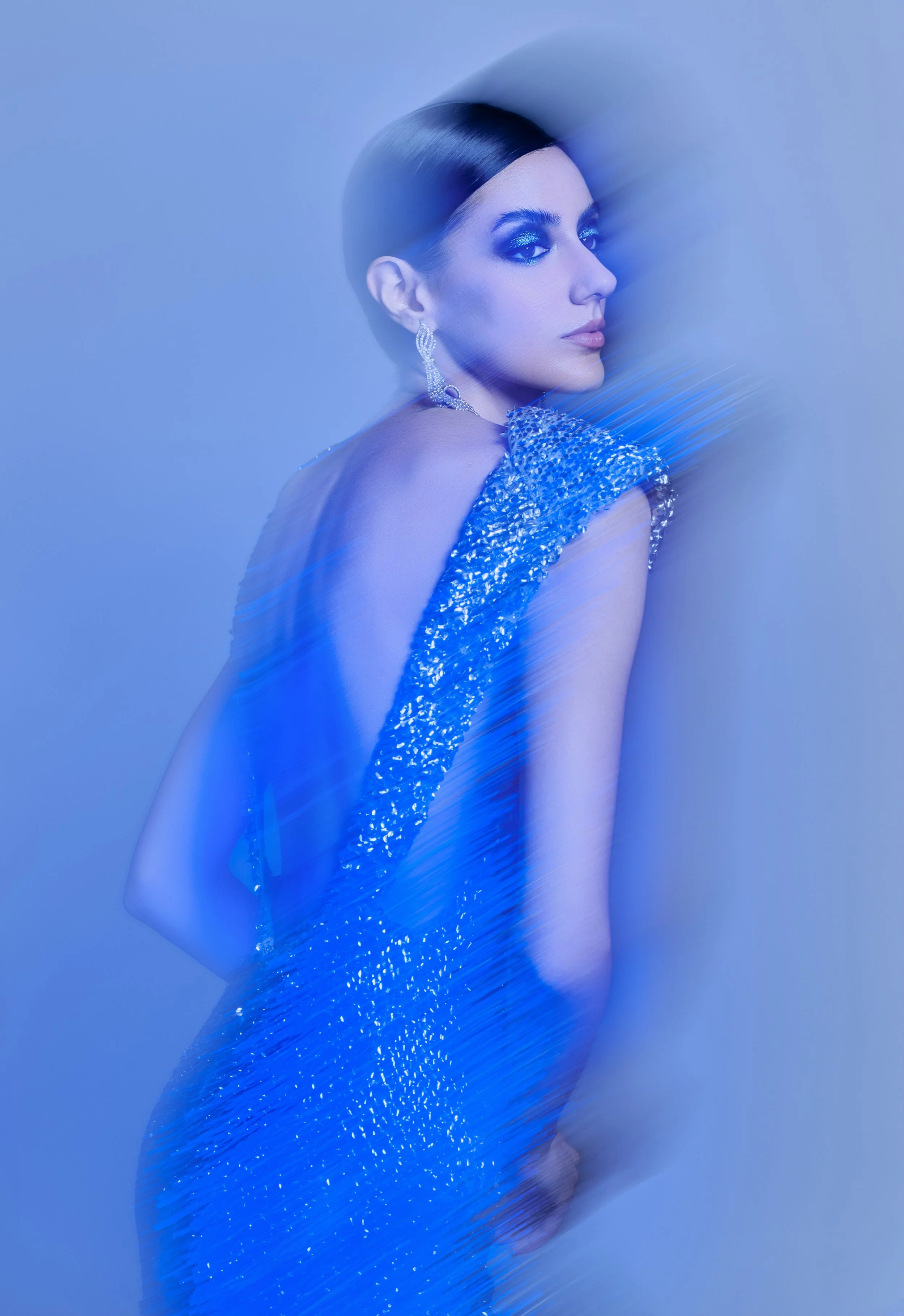 A woman with dark hair styled sleekly, wearing a sparkling blue dress and large earrings, posing against a blue background with motion blur effect.