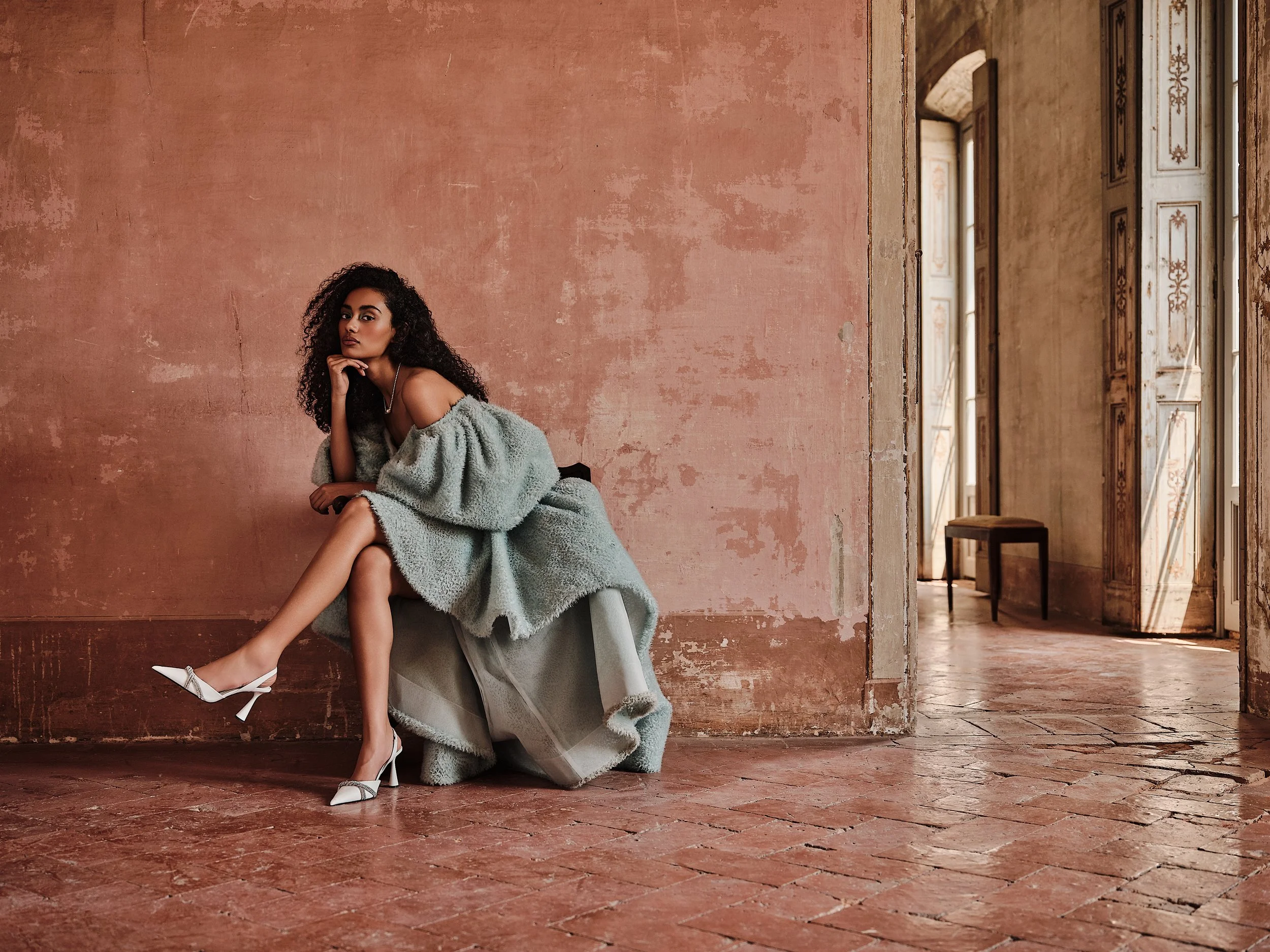A woman with curly black hair sitting on a gray chair in a spacious room with textured pink and beige walls and large open wooden doors, wearing a gray plush off-the-shoulder dress and white high heels.