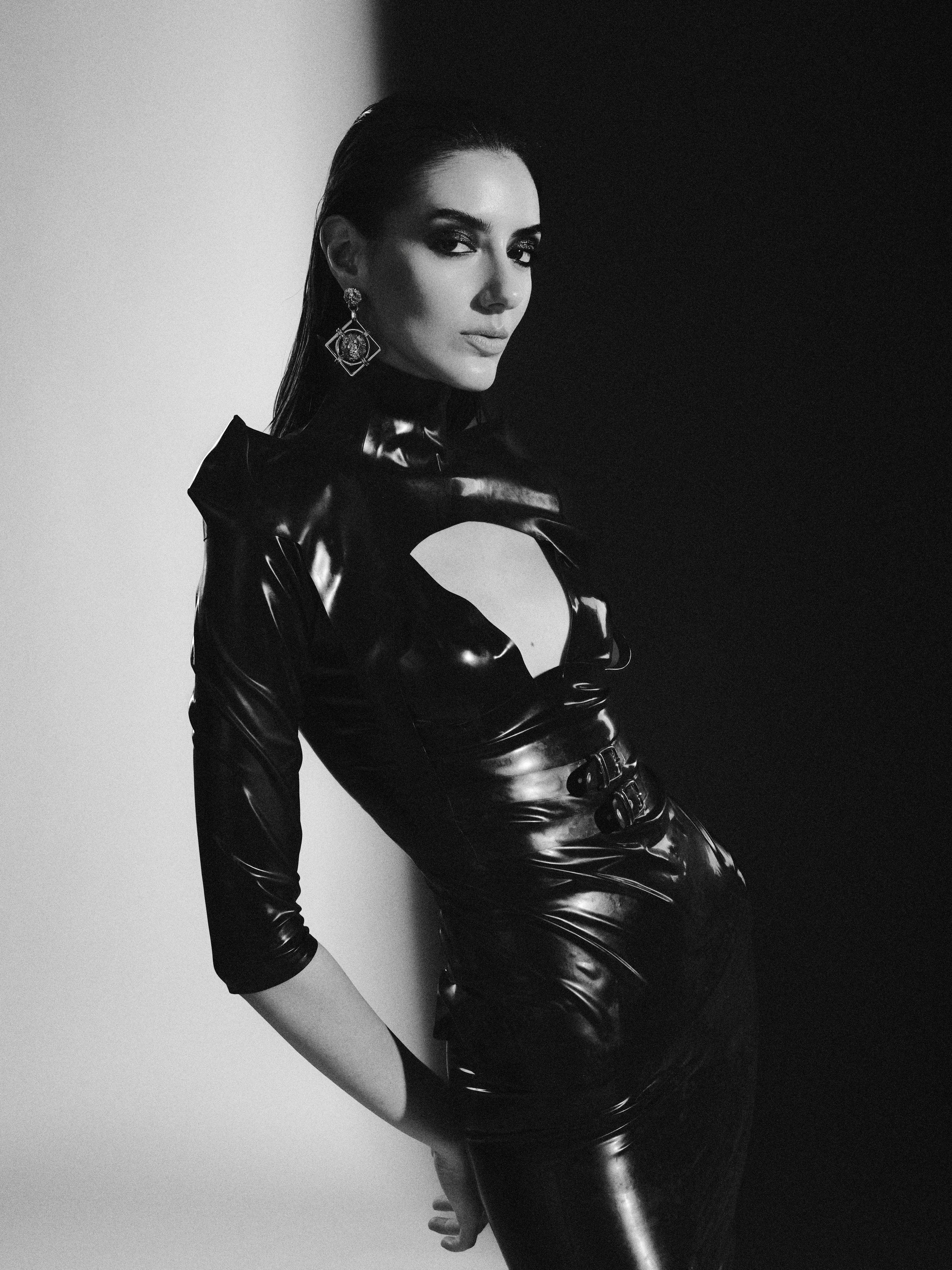 A woman with dark hair, wearing earrings and a shiny, black, fitted dress with cutouts, poses against a background split into white and black halves.