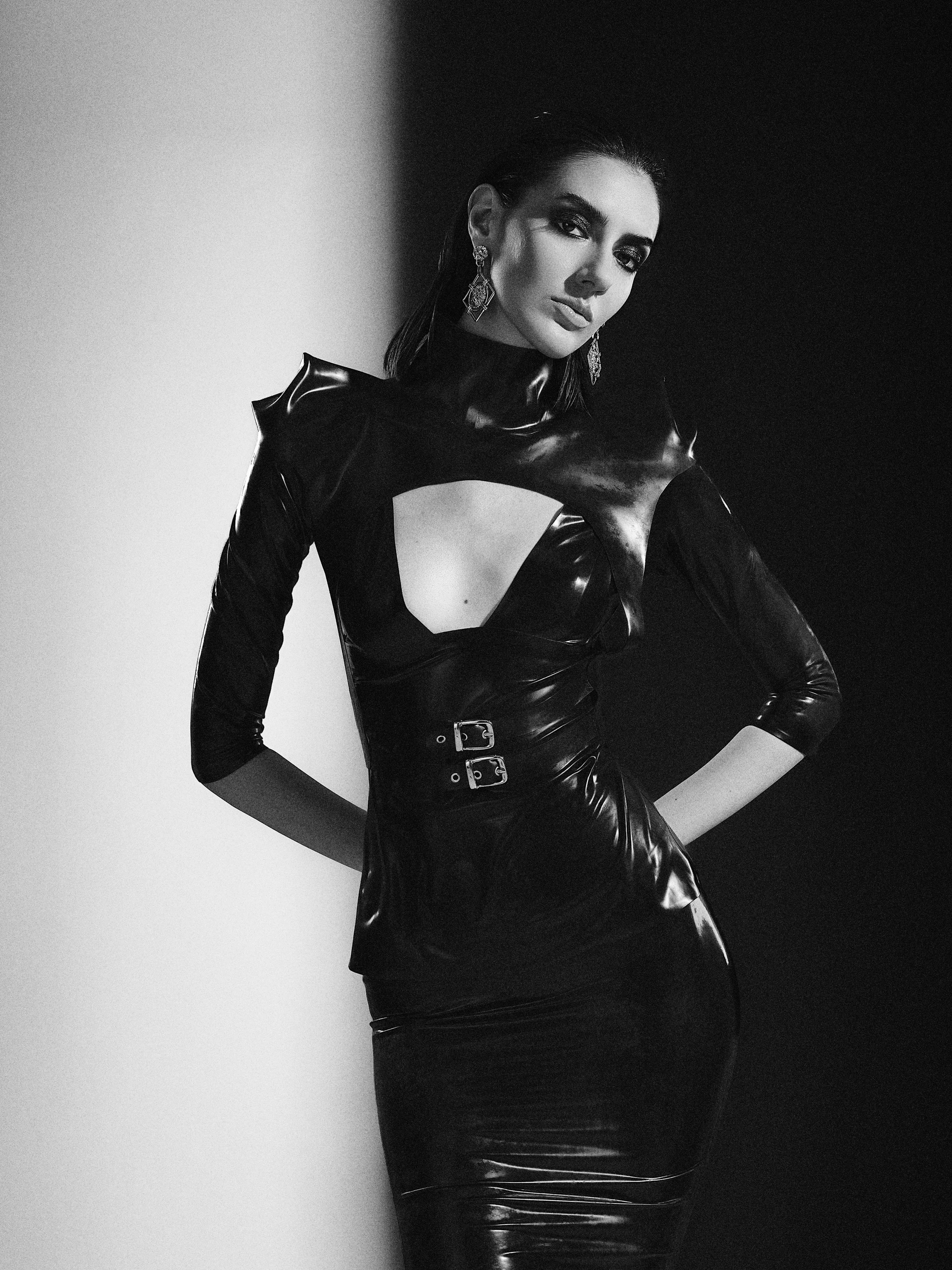 A woman with dark hair, wearing a shiny black latex dress with cut-out details and buckles, standing against contrasting black and white background, accessorized with dangling earrings.
