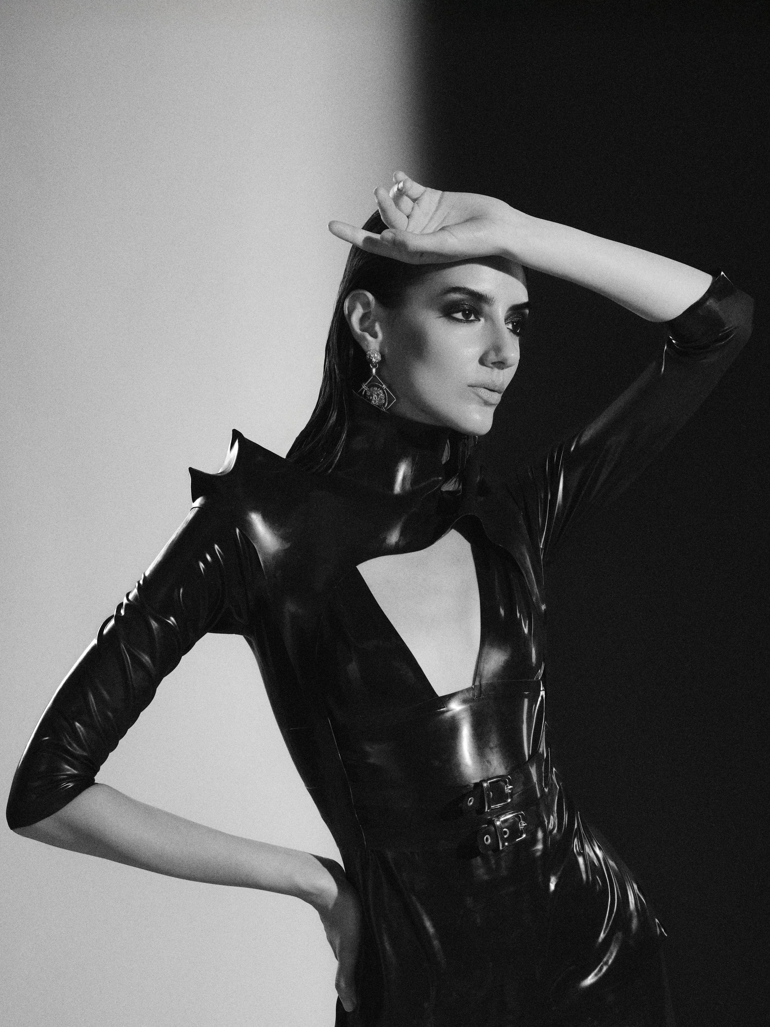 A woman with dark hair and makeup wearing a shiny black latex dress with cutout design, holding one arm over her face, in front of a two-tone background.