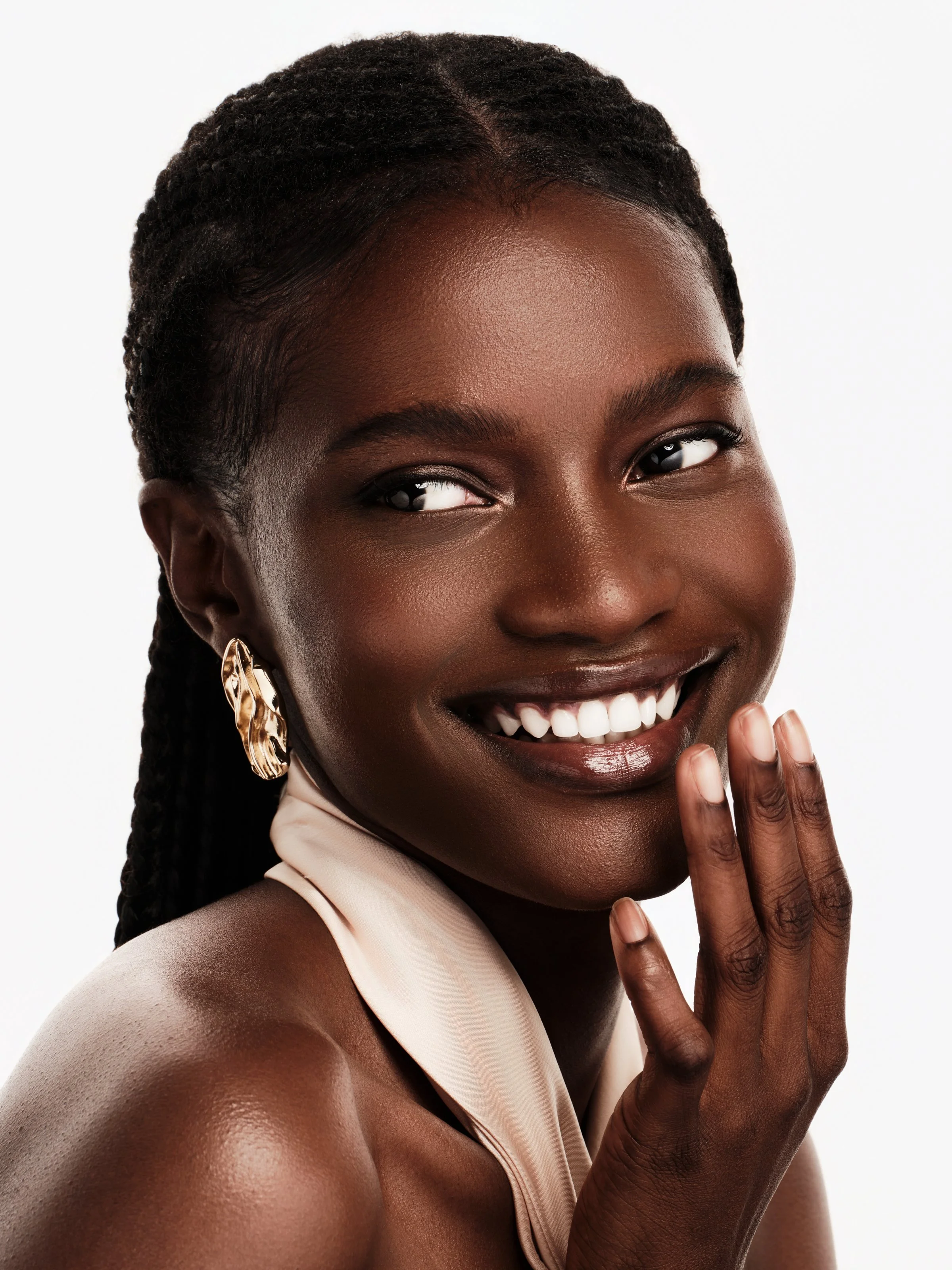 Close-up of a smiling Black woman with braided hair, wearing gold earrings, and a beige sleeveless top, touching her face gently.