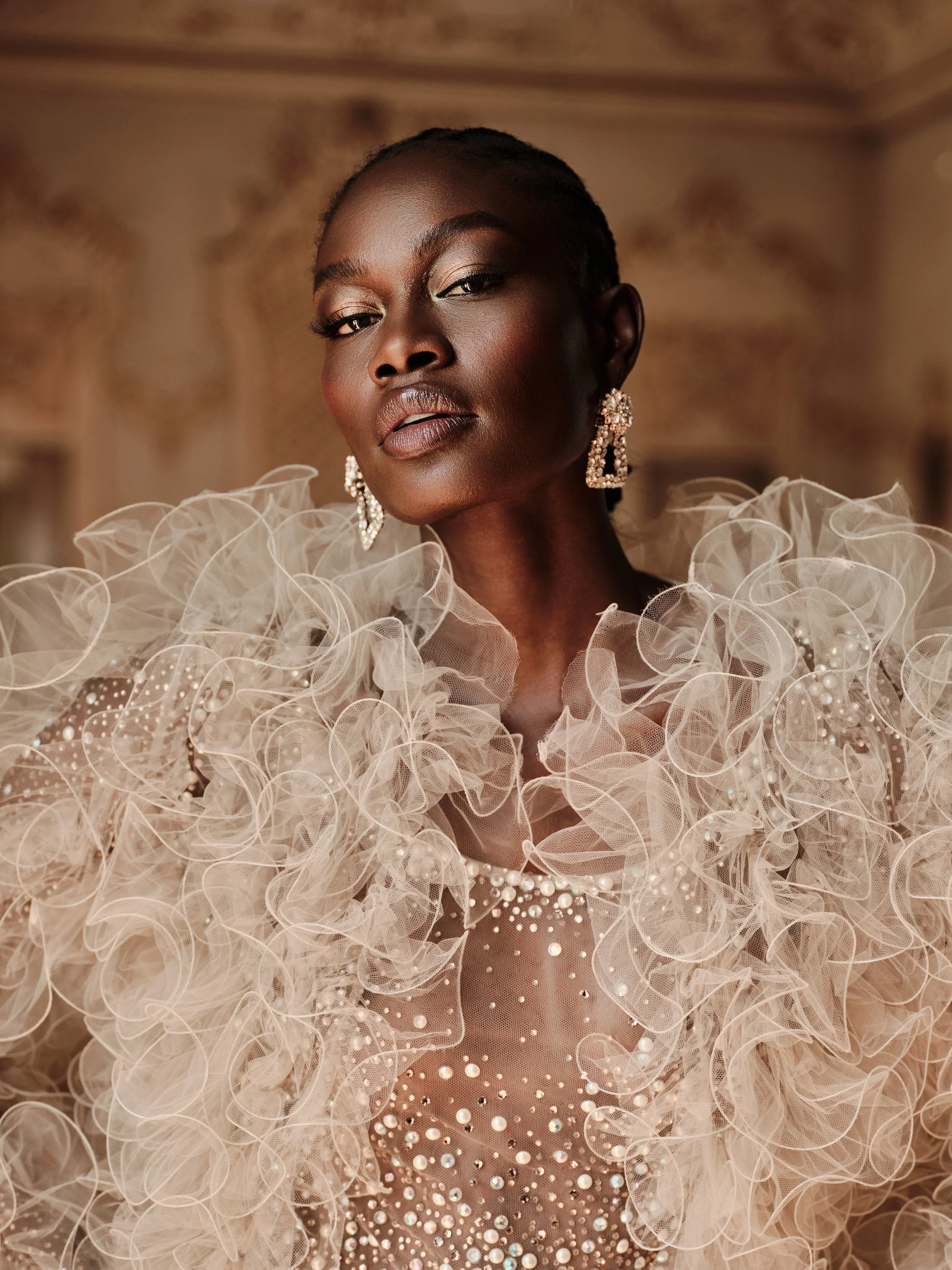 A woman with dark skin and short hair wearing a detailed, ruffled, pearl-embellished gown and matching earrings, posing elegantly indoors.