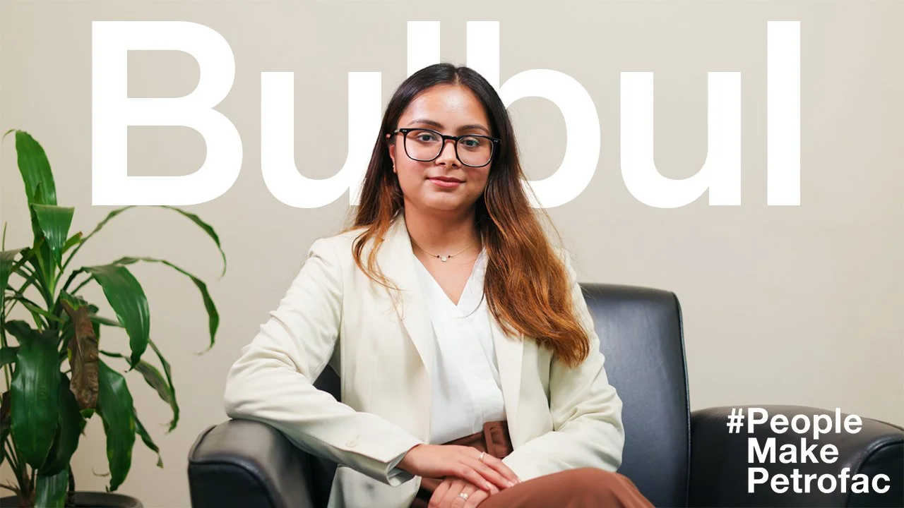 A young woman with glasses and brown hair sitting on a black chair in front of a large sign that says 'Bubol'. She is wearing a white blazer and a white blouse, with a plant on her left side. The text '#People Make Petrofac' is at the bottom right corner.