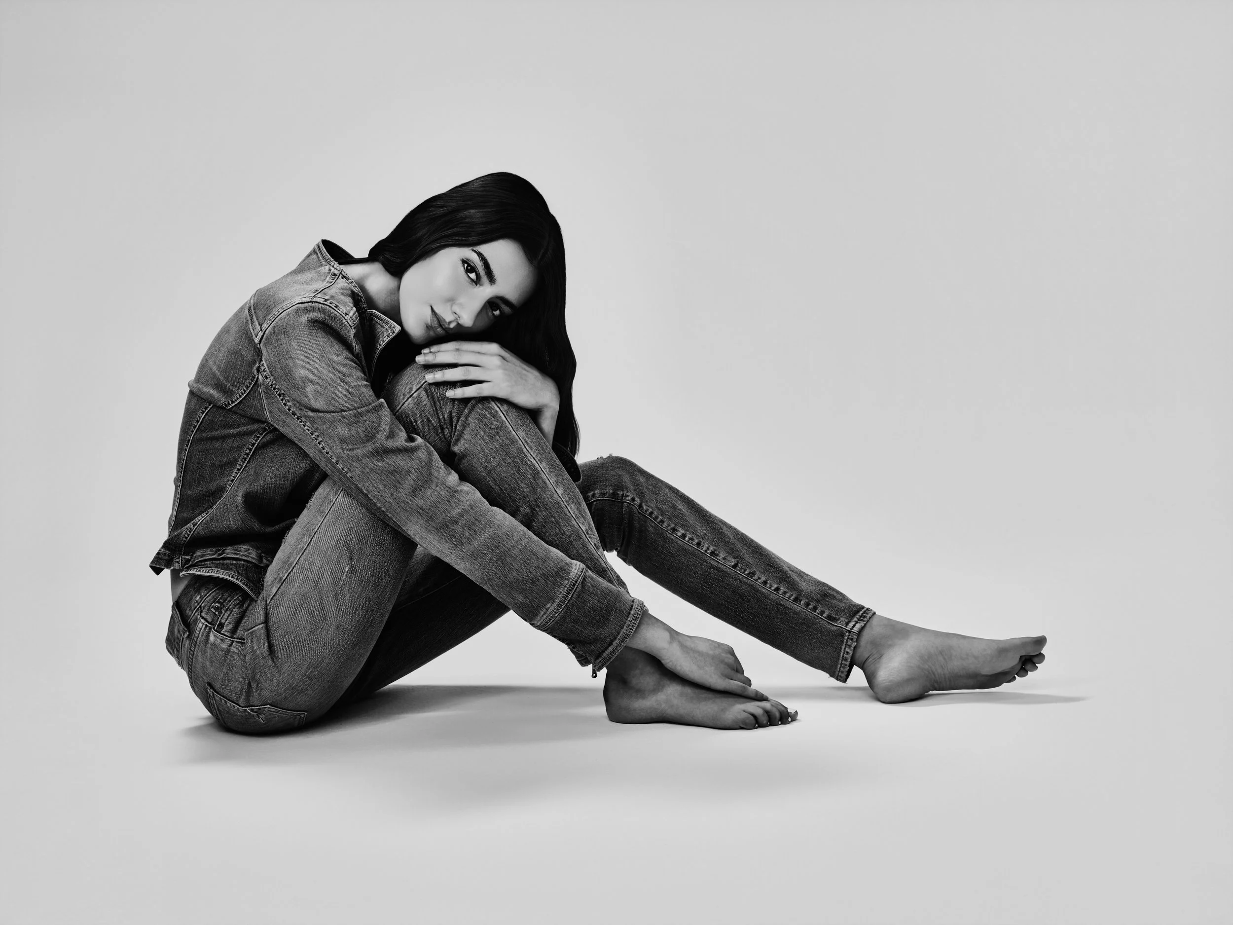 Black and white photo of a woman sitting on the floor in a relaxed pose, wearing denim jeans and a denim jacket, with long dark hair and a calm expression.