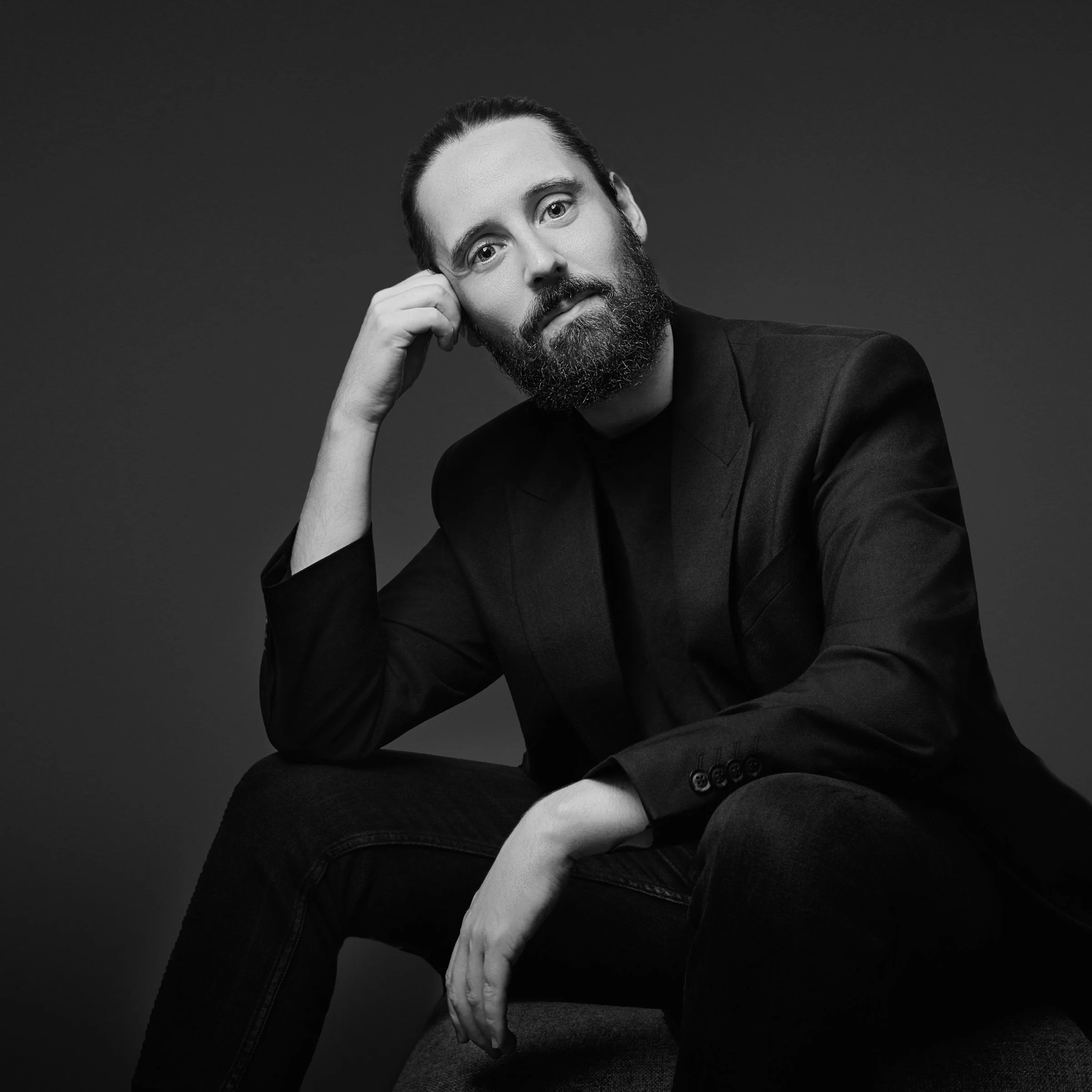 Black and white portrait of Darren Hill, a creative director with a beard, wearing a blazer and sitting on a chair with a dark background.