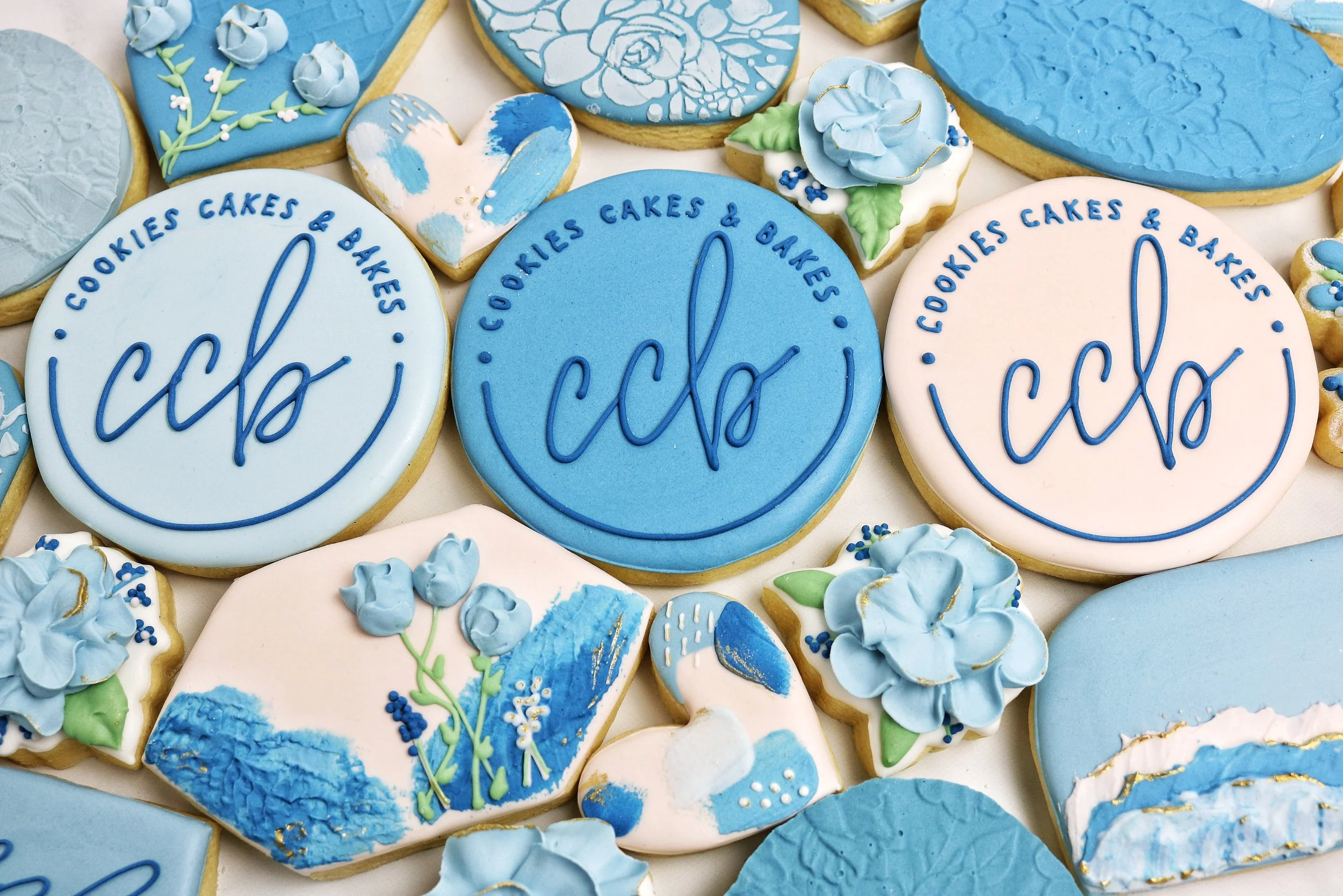 Sugar cookies decorated with royal icing to match the Cookies Cakes & Bakes logo.  The cookies show script "ccb" on the cookies.  Foral cookies are around the logo cookies.