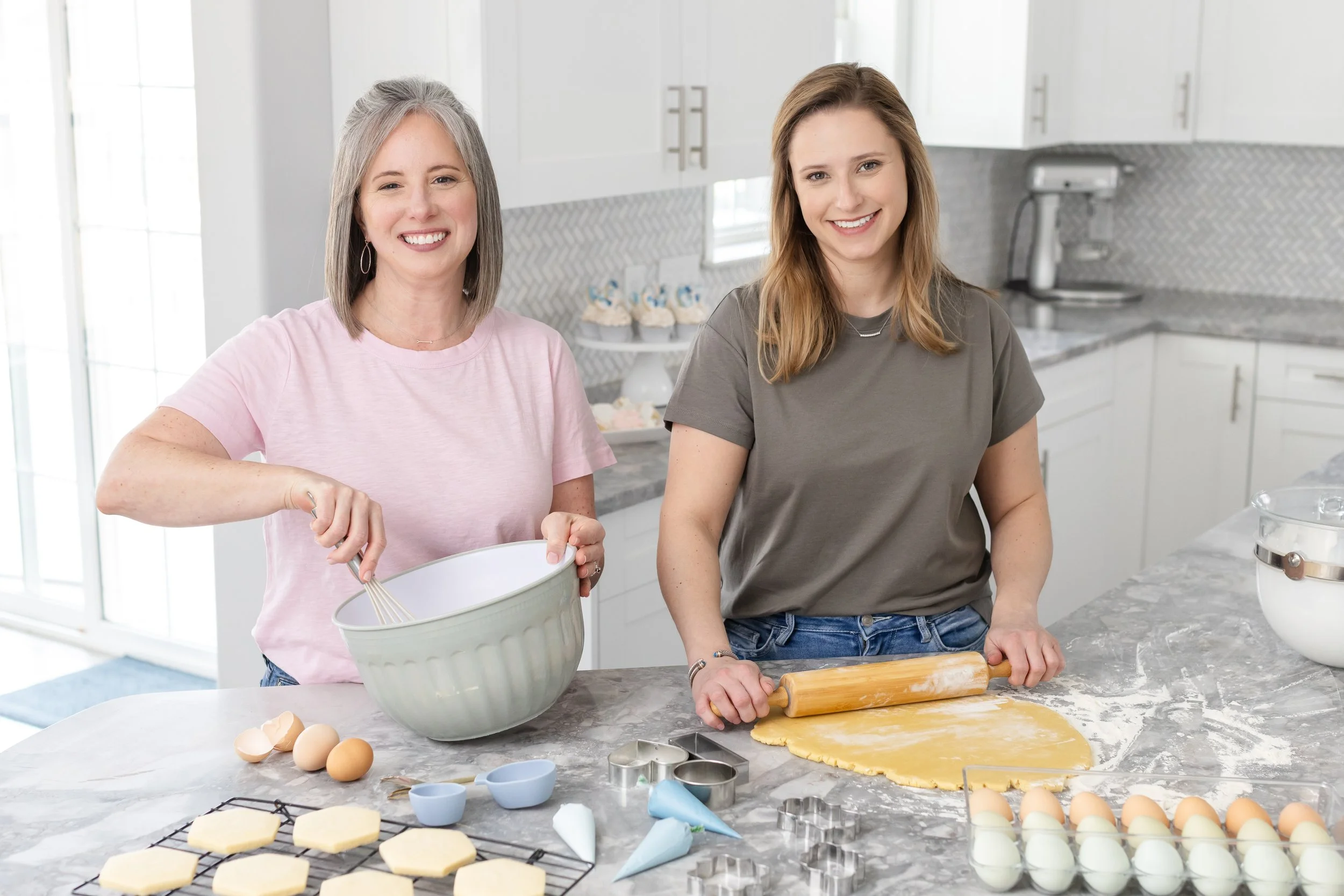 2 women smiling in a bright, modern kitchen baking together.  One woman whisks ingredients while the other rolls out cookie dough.  There are many baking items on the counter around them.