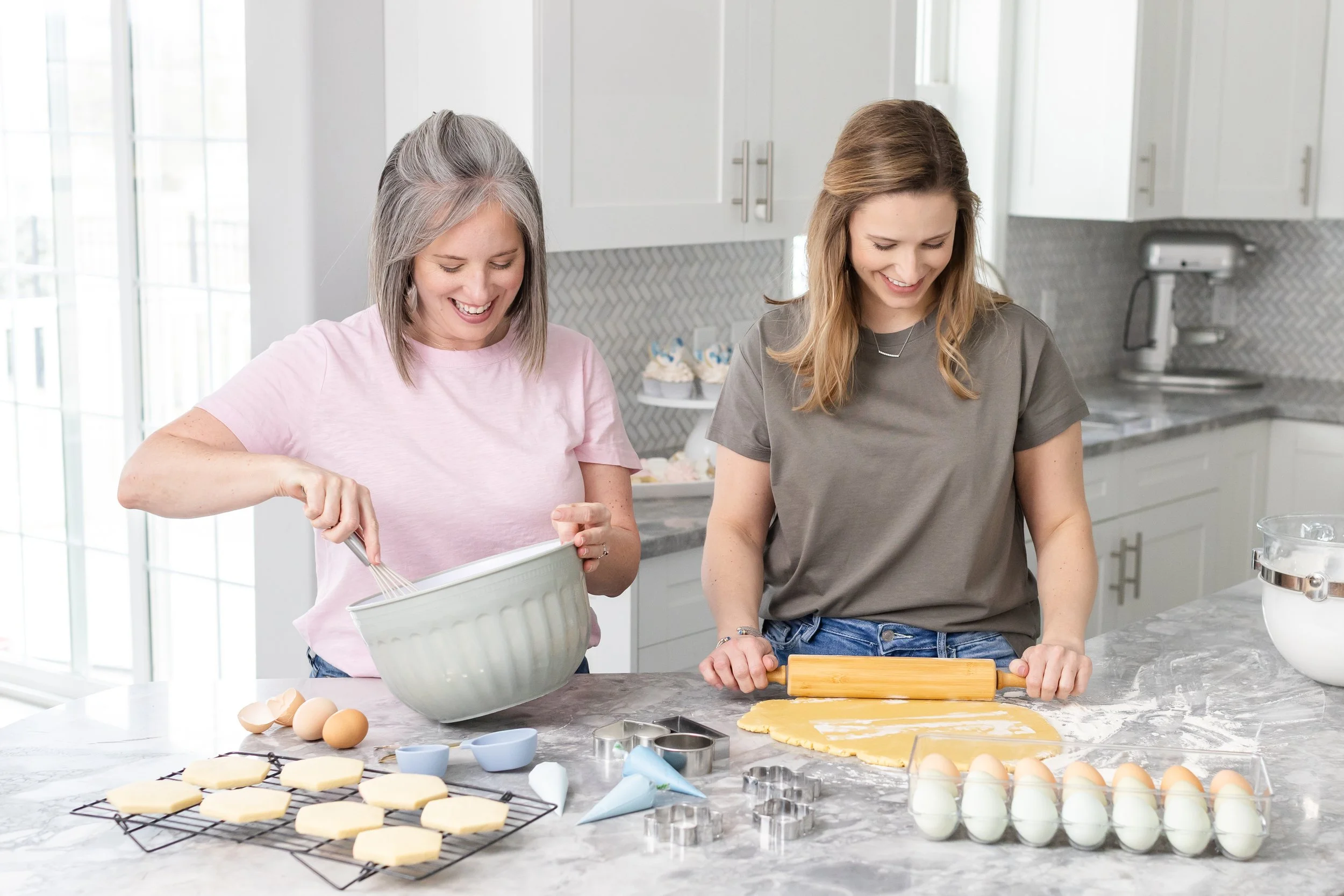 2 women laughing in a bright, modern kitchen baking together.  One woman whisks ingredients while the other rolls out cookie dough.  There are many baking items on the counter around them.