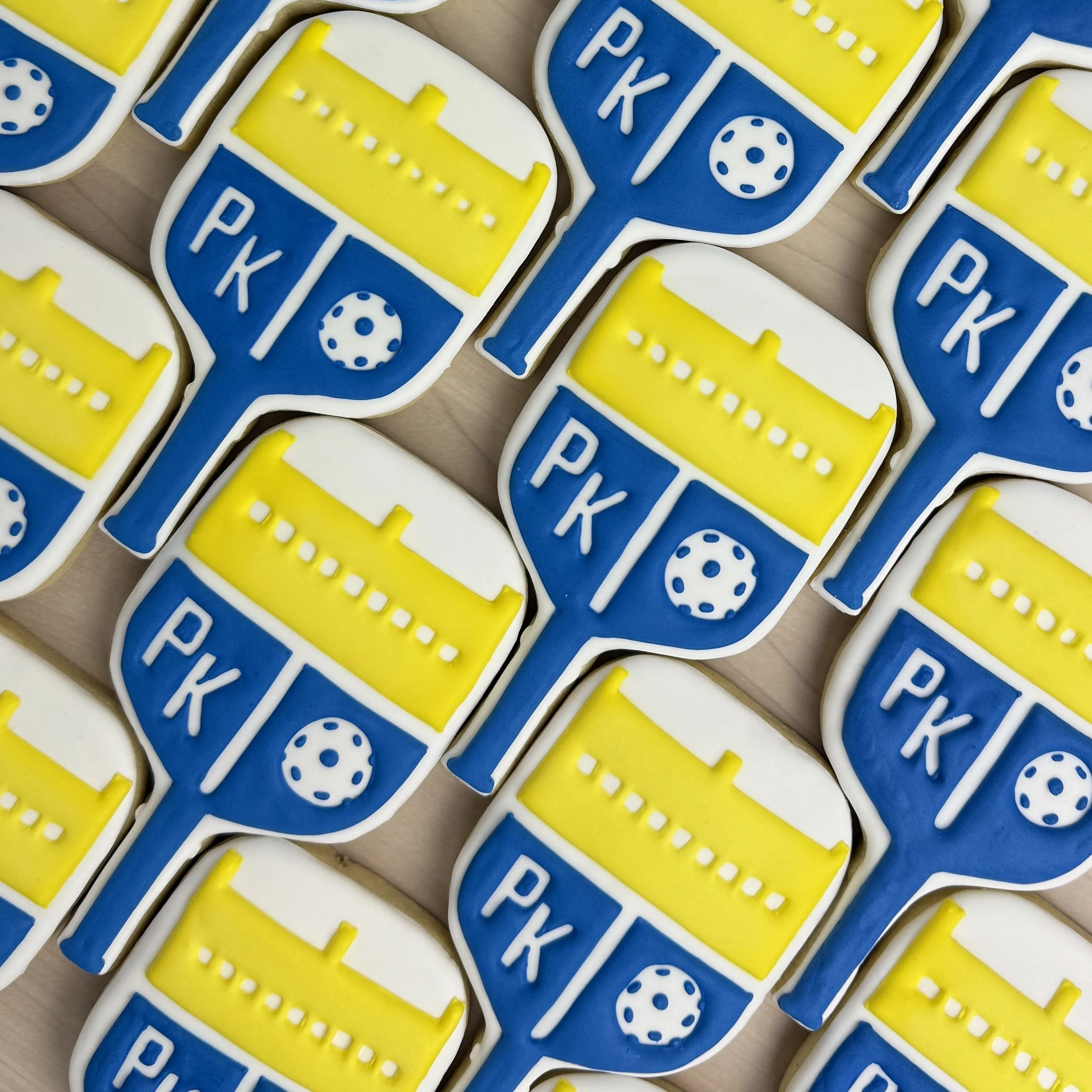 Royal icing decorated sugar cookies in the shape of pickleball paddle.  Cookies have blue icing with PK and ball in white, and yellow top half of the paddle.