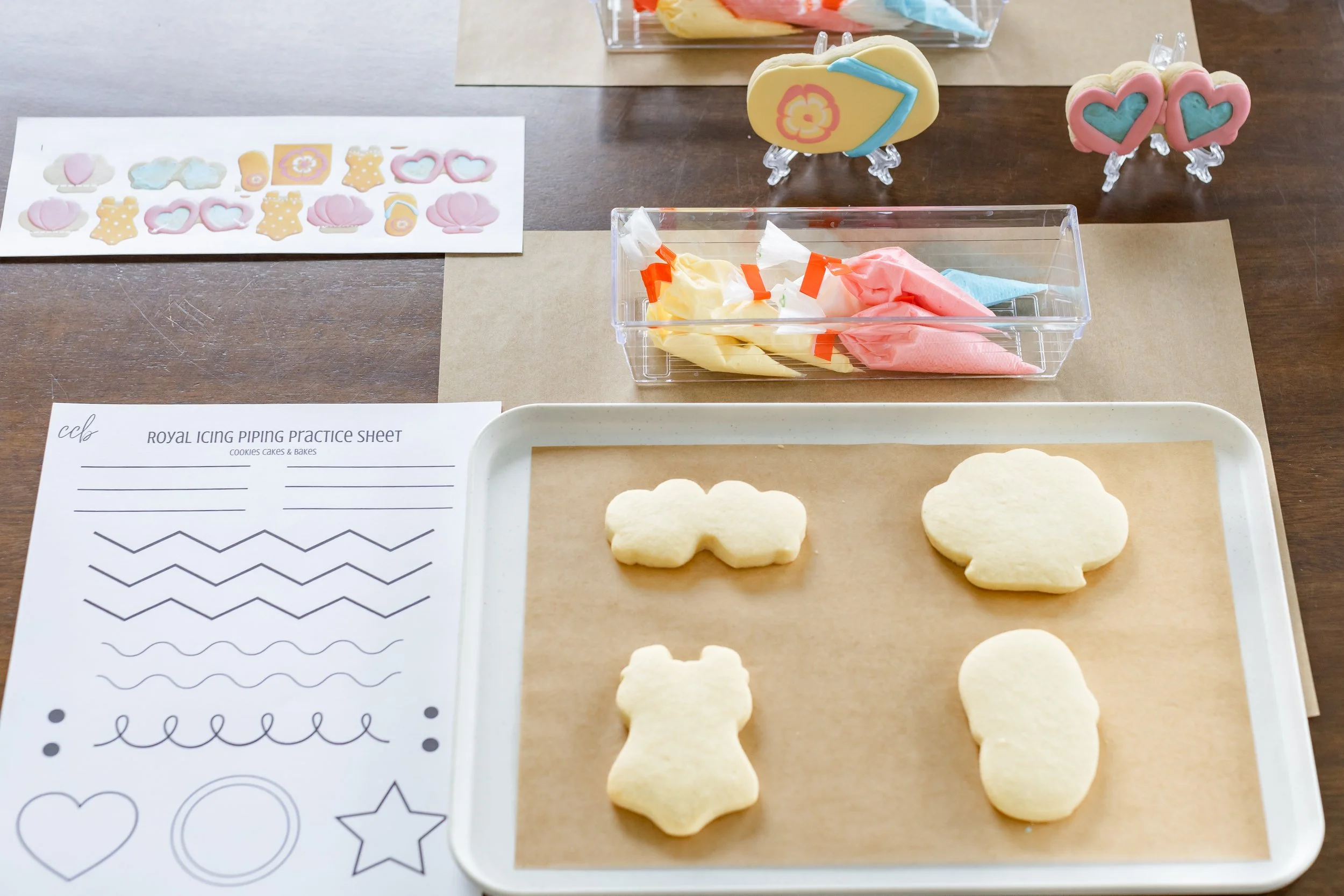 A wooden table set up for a cookie decorating class.  On the table is a tray with plain cookies, bags of icing, a practice sheet for piping, and a few beach themed, decorated cookies.