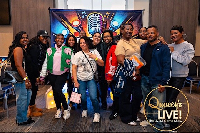 Group of people posing in front of a colorful backdrop with a large microphone and music notes, at a show called 'Quacey's LIVE! The LIVEST Game Show'.
