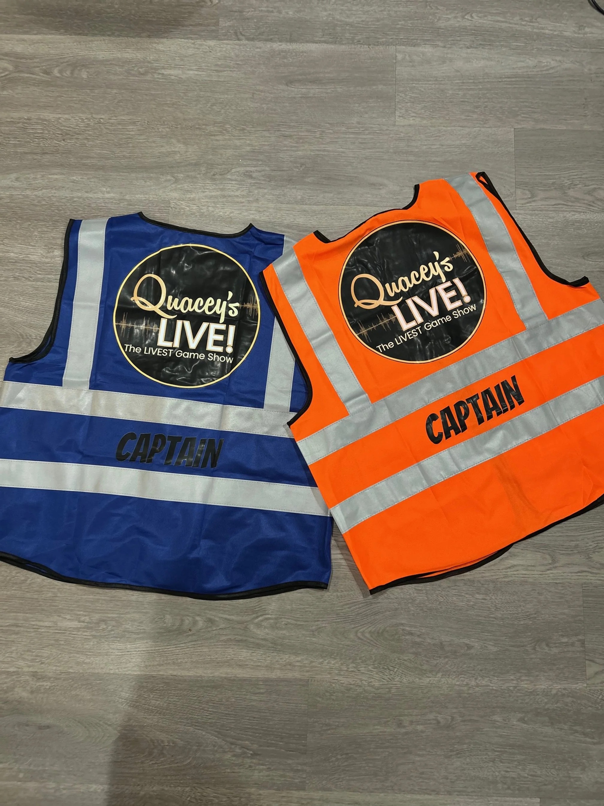 Two reflective safety vests, one blue and one orange, lying on a wooden floor. Both have a circular logo on the front reading "Quacey's LIVE! The LIVEST Game Show," and the word "CAPTAIN" printed in bold letters across the middle.