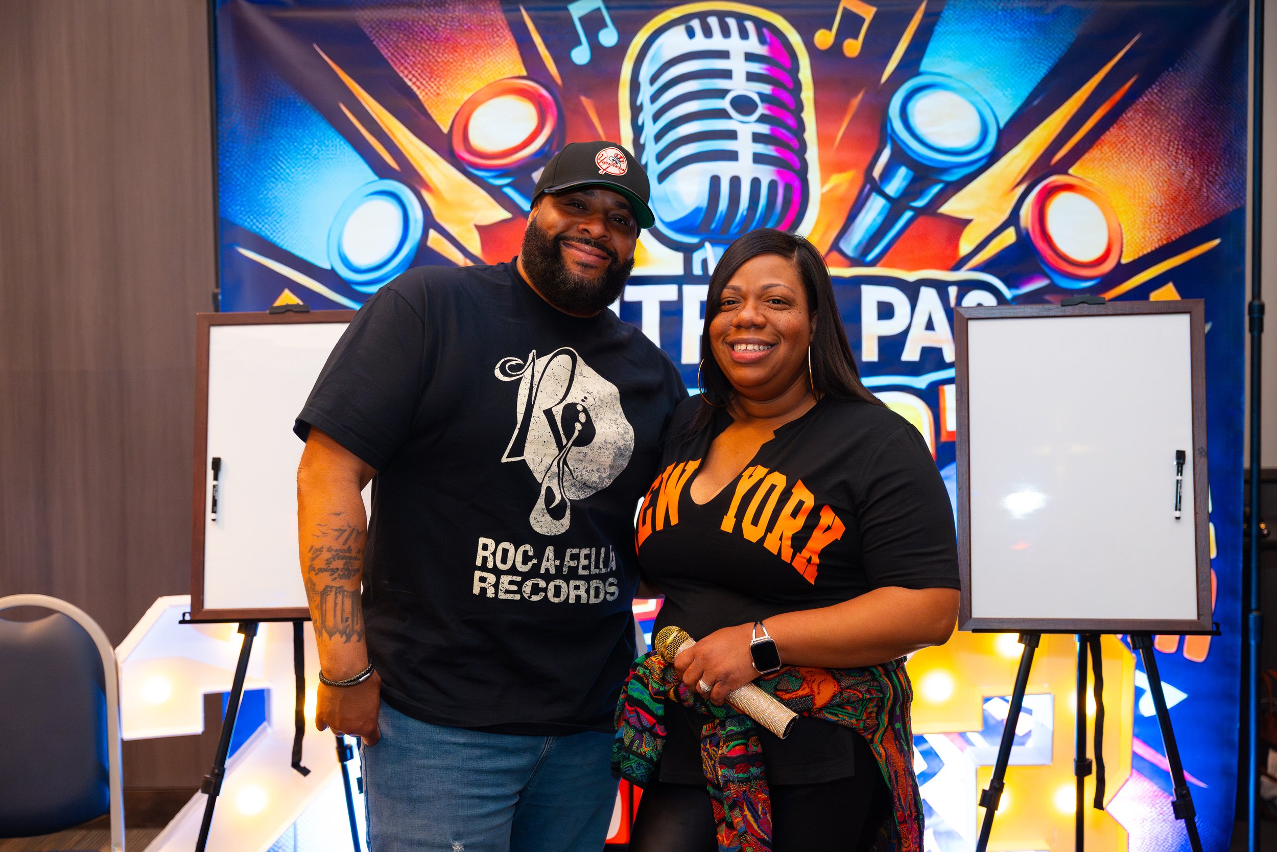 Two people smiling at a microphone, standing together indoors with a colorful music-themed backdrop.