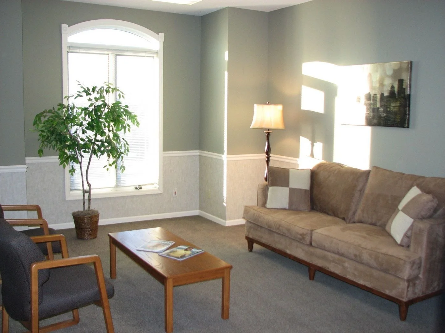 A living room with a beige sofa, two throw pillows, a wooden coffee table with magazines, a gray armchair with wooden armrests, a floor lamp, a large potted plant, a window with closed blinds, and a piece of art depicting a city skyline.
