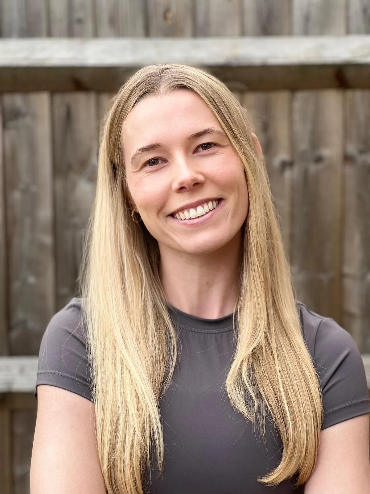 A young woman with long blonde hair, smiling while standing outdoors in front of a wooden fence.