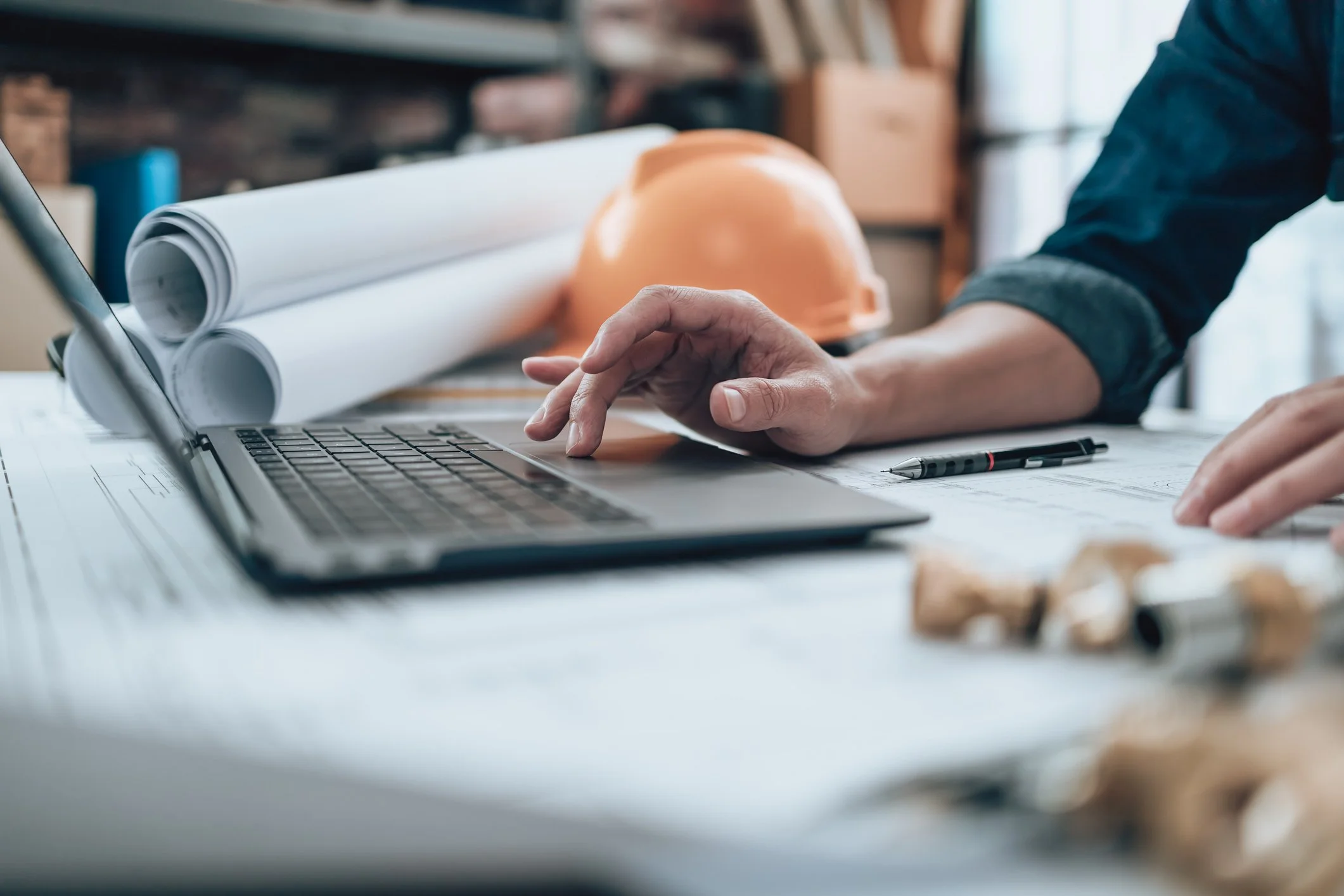 A person working on a laptop at a desk with blueprints, a hard hat, and architectural plans nearby.