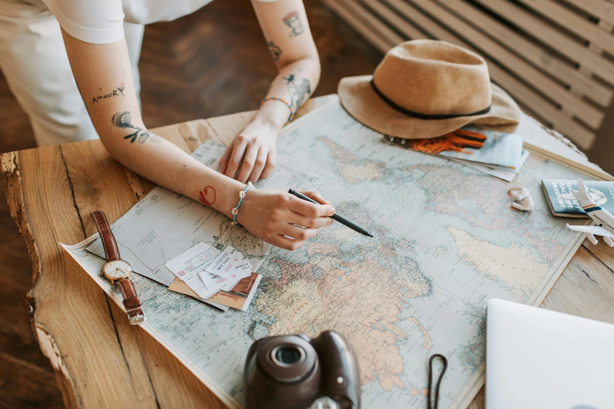 Person planning a trip with a world map, travel tickets, a camera, and accessories on a rustic wooden table.