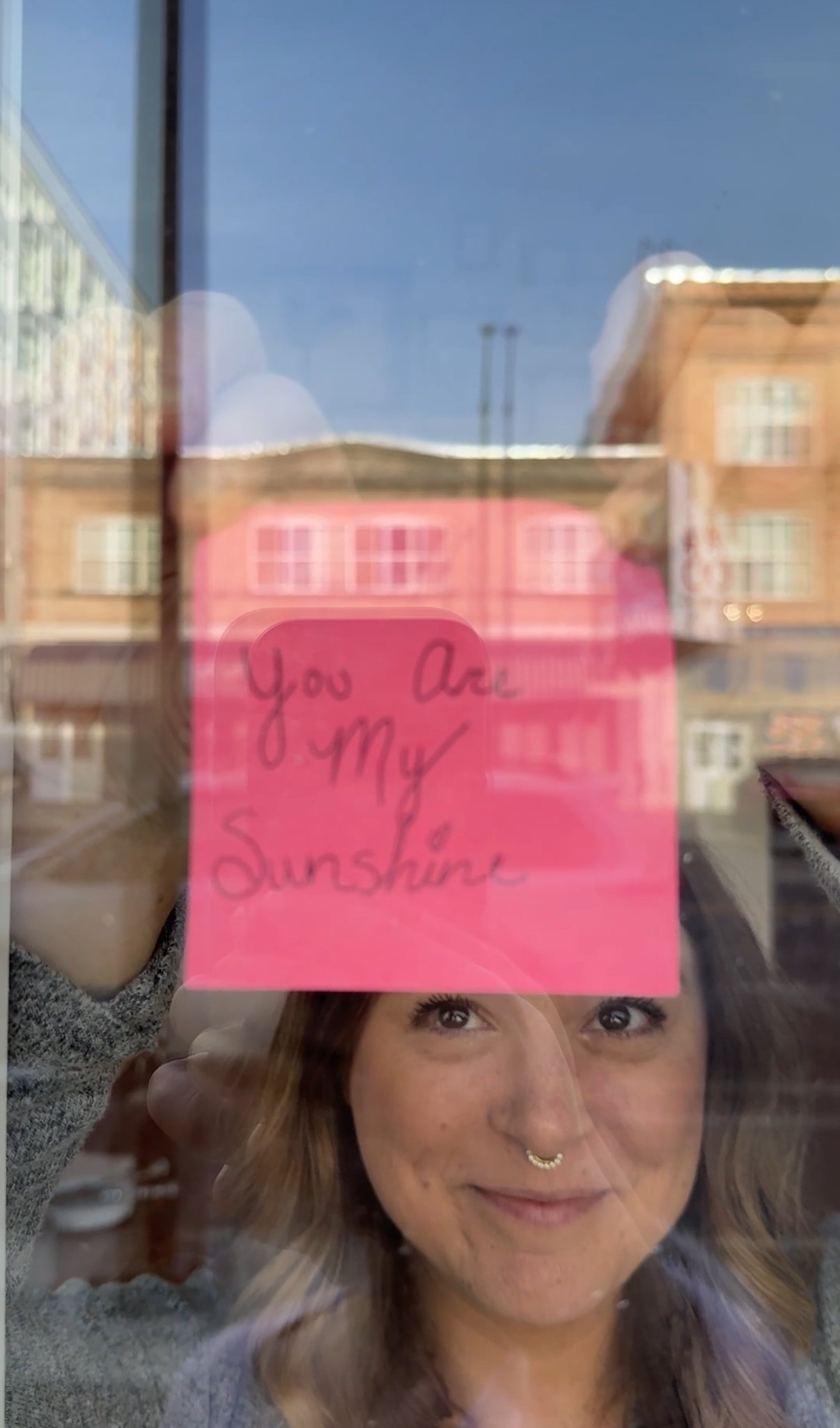A woman with light brown hair and a septum piercing holding a pink note with the handwritten message 'You Are My Sunshine' on a window, with buildings and a clear blue sky reflected in the glass.