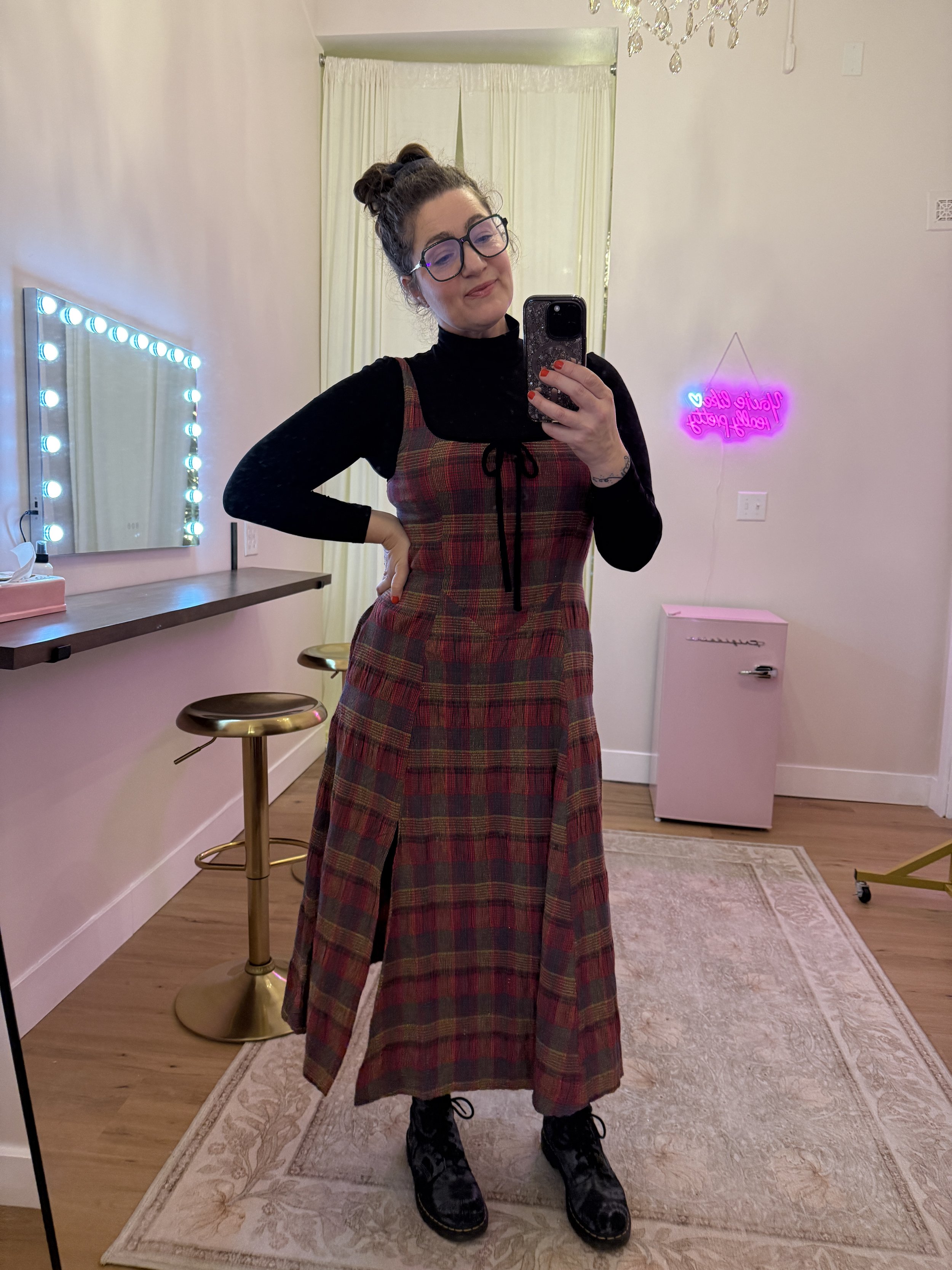 A woman taking a mirror selfie in a room with pink walls and a hardwood floor, wearing black glasses, a black turtleneck, and a plaid dress, with her hair in a bun and sneakers.