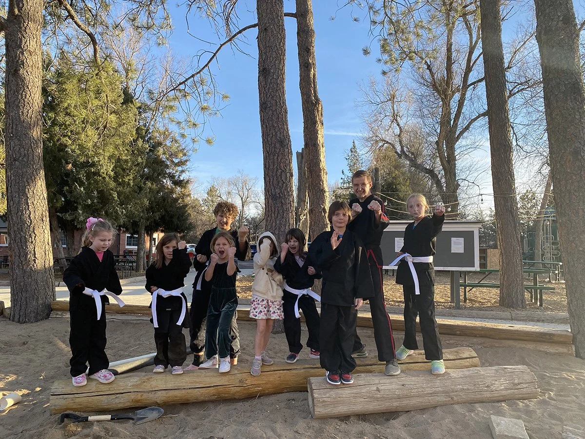 Group of children and a young adult posing on a wooden balance beam in an outdoor park, some in martial arts uniforms, making fighting stances, with trees and a community building in the background.