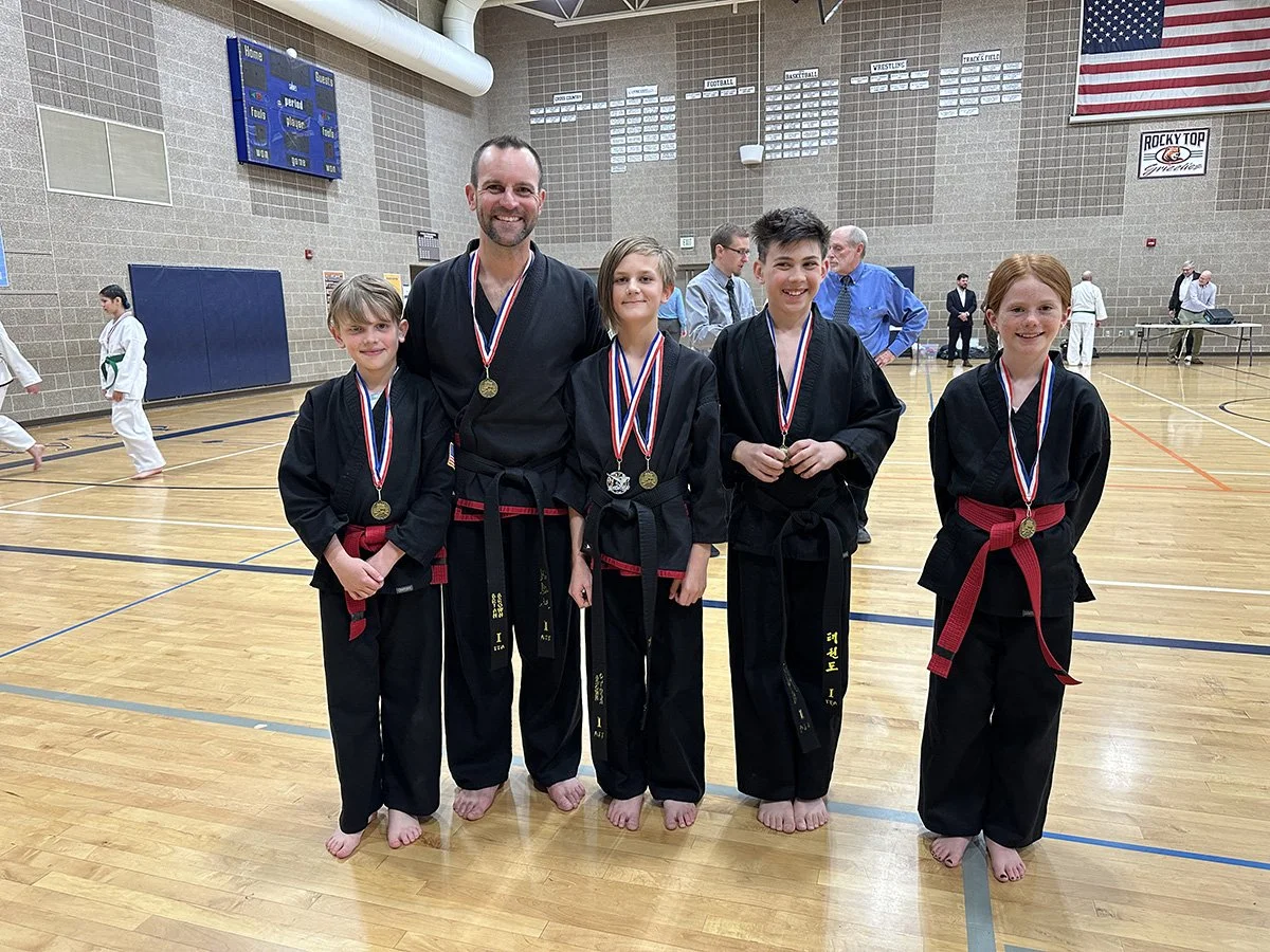 A group of five martial artists, four children and one adult, standing in a gymnasium with medals around their necks. They are all wearing black uniforms with red belts and are smiling at the camera. In the background, other martial artists and offic