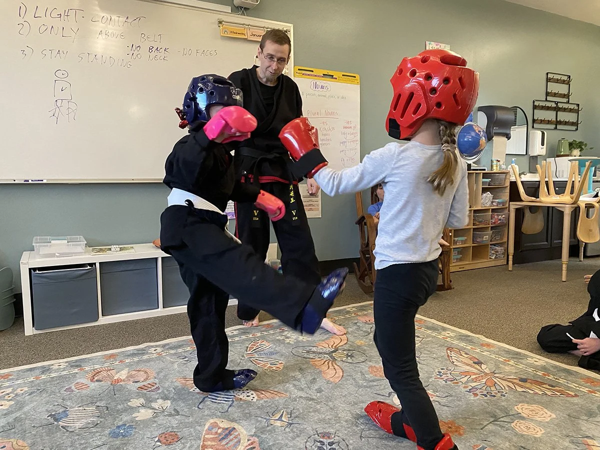Two children in martial arts uniforms with helmets and gloves sparring in a classroom. An instructor watches them, and teachers' notes are written on a whiteboard in the background.