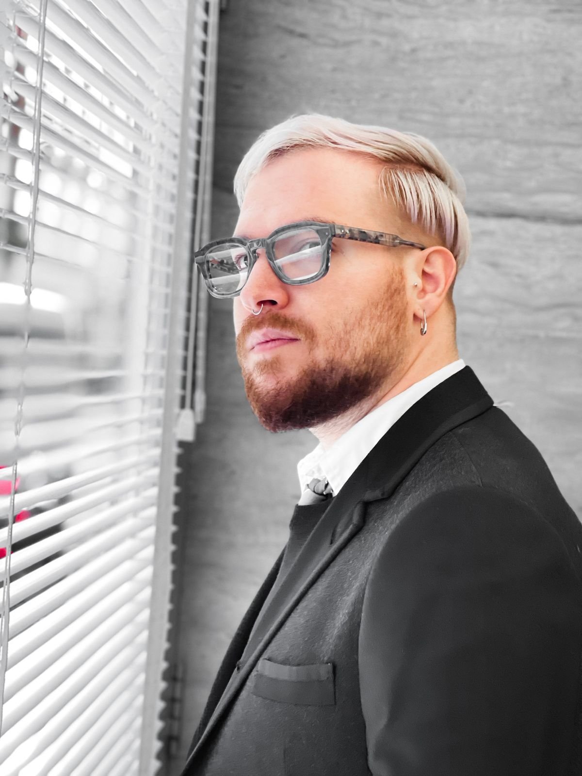 George, wearing a black suit and glasses, looking out through window blinds with soft natural light on his face.