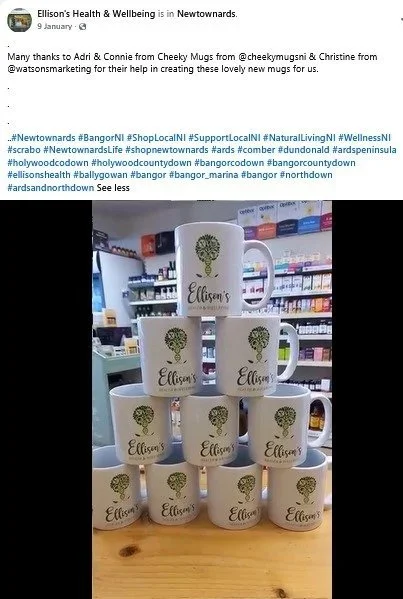 Stacked white ceramic mugs with a green tree logo and the name Ellen's on a wooden table, with shelves of products in the background.
