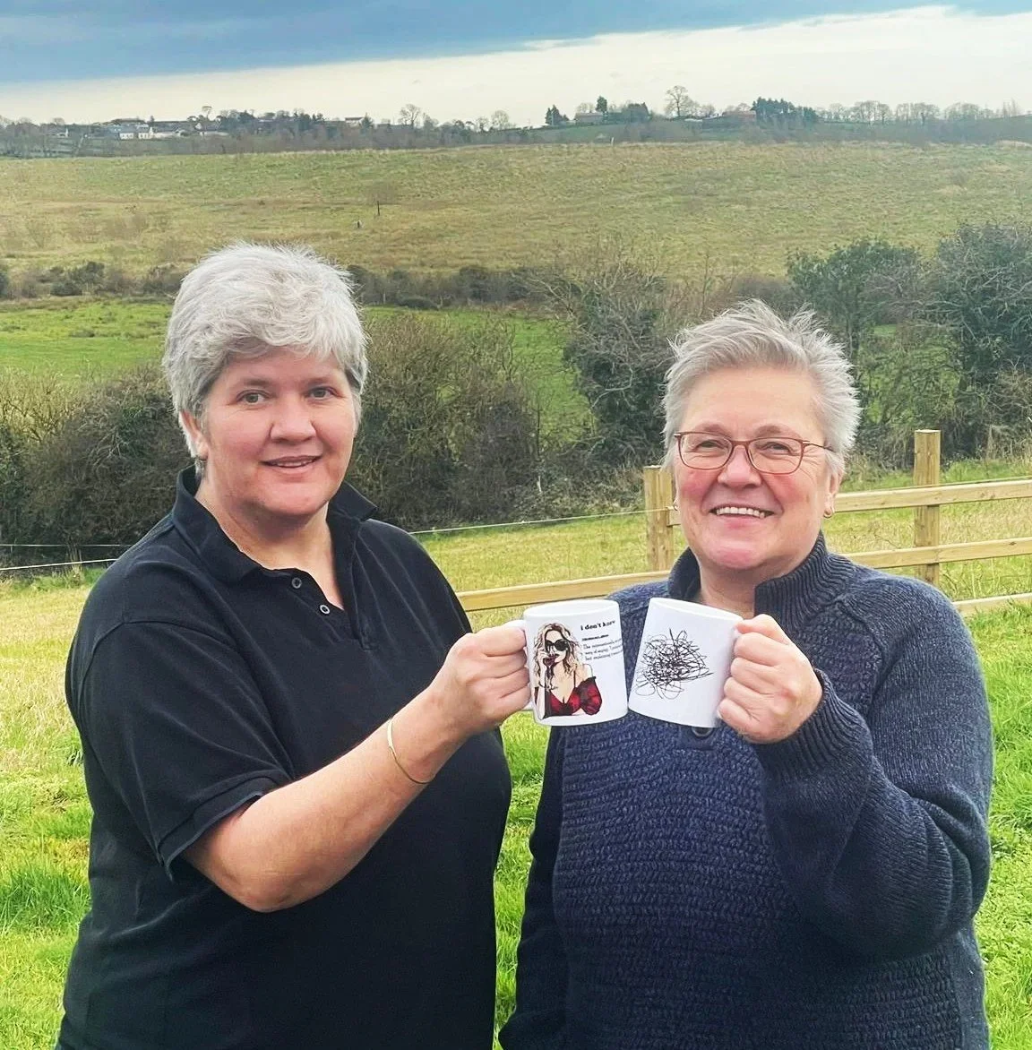 Two smiling women holding coffee mugs outdoors in a grassy field with rolling hills and cloudy sky in the background.