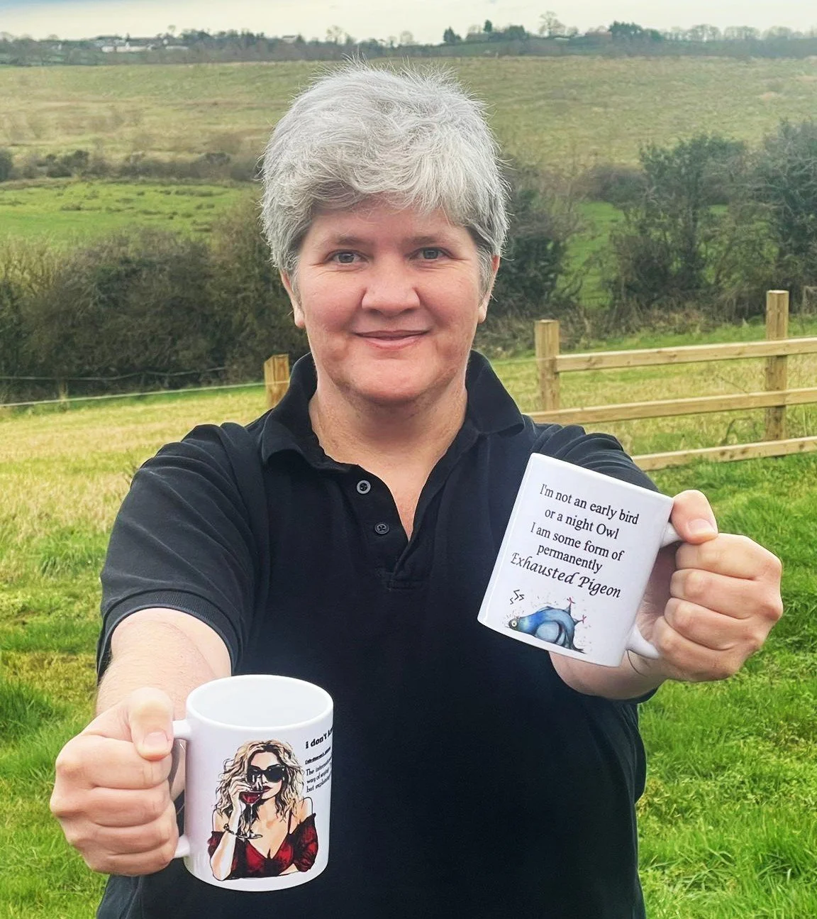 A woman with short gray hair outdoors holding two mugs with humorous messages and images. The background shows a grassy field, trees, and rolling hills.