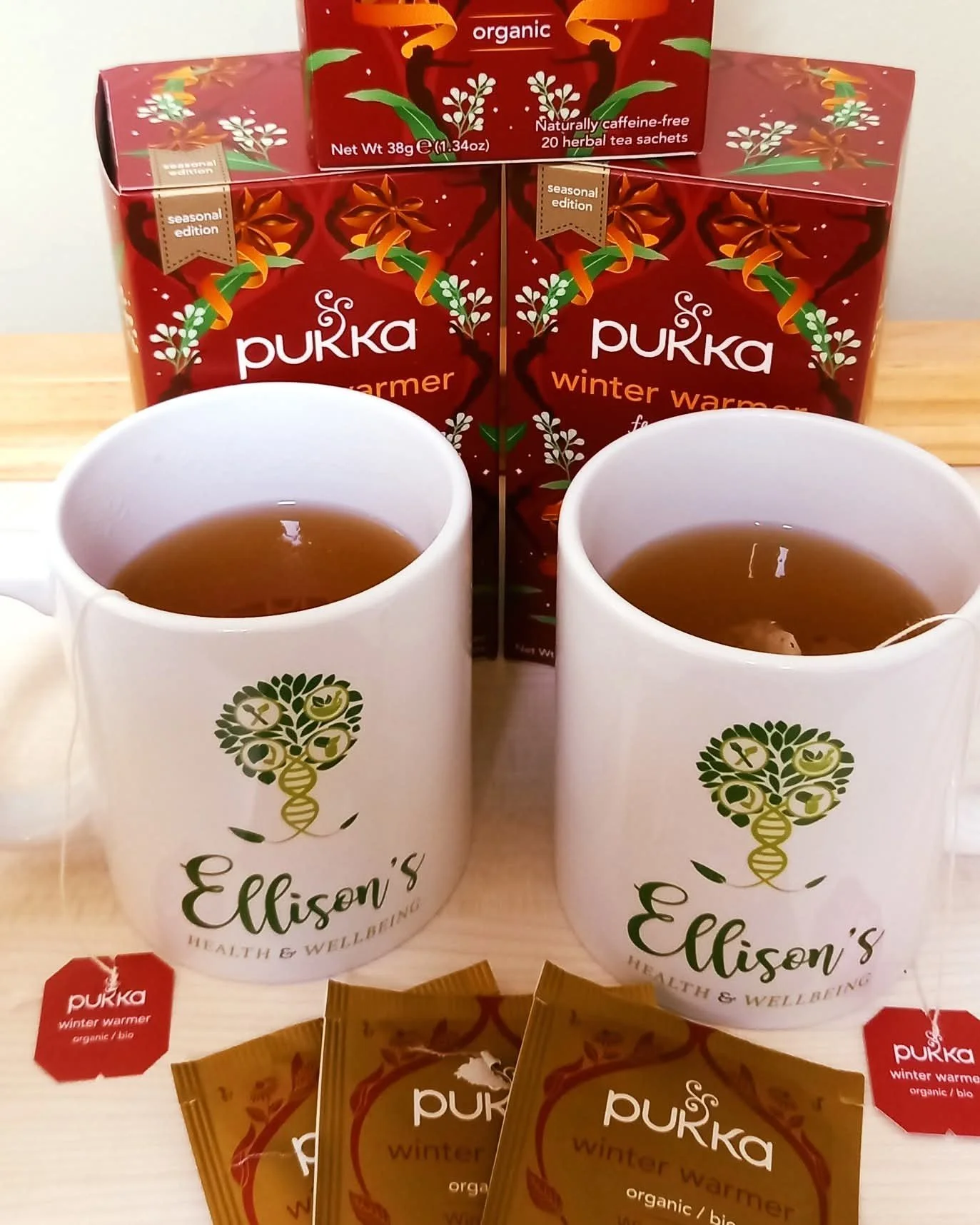 Two white mugs with green logo and text reading 'Ellison's health &wellbeing,' filled with tea, in front of two red boxes of Pukka winter warmer herbal tea with colorful autumn-themed design. Several tea sachets and packets are also visible.
