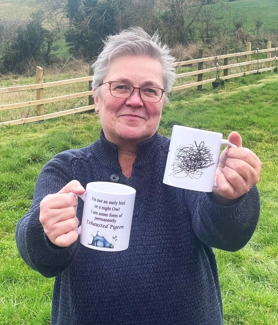 A woman with short gray hair, glasses, and a navy knit sweater smiling outdoors in a grassy area with a wooden fence behind her, holding two coffee mugs. One mug has a tangled scribble design; the other has a humorous message about being a permanently exhausted pigeon.