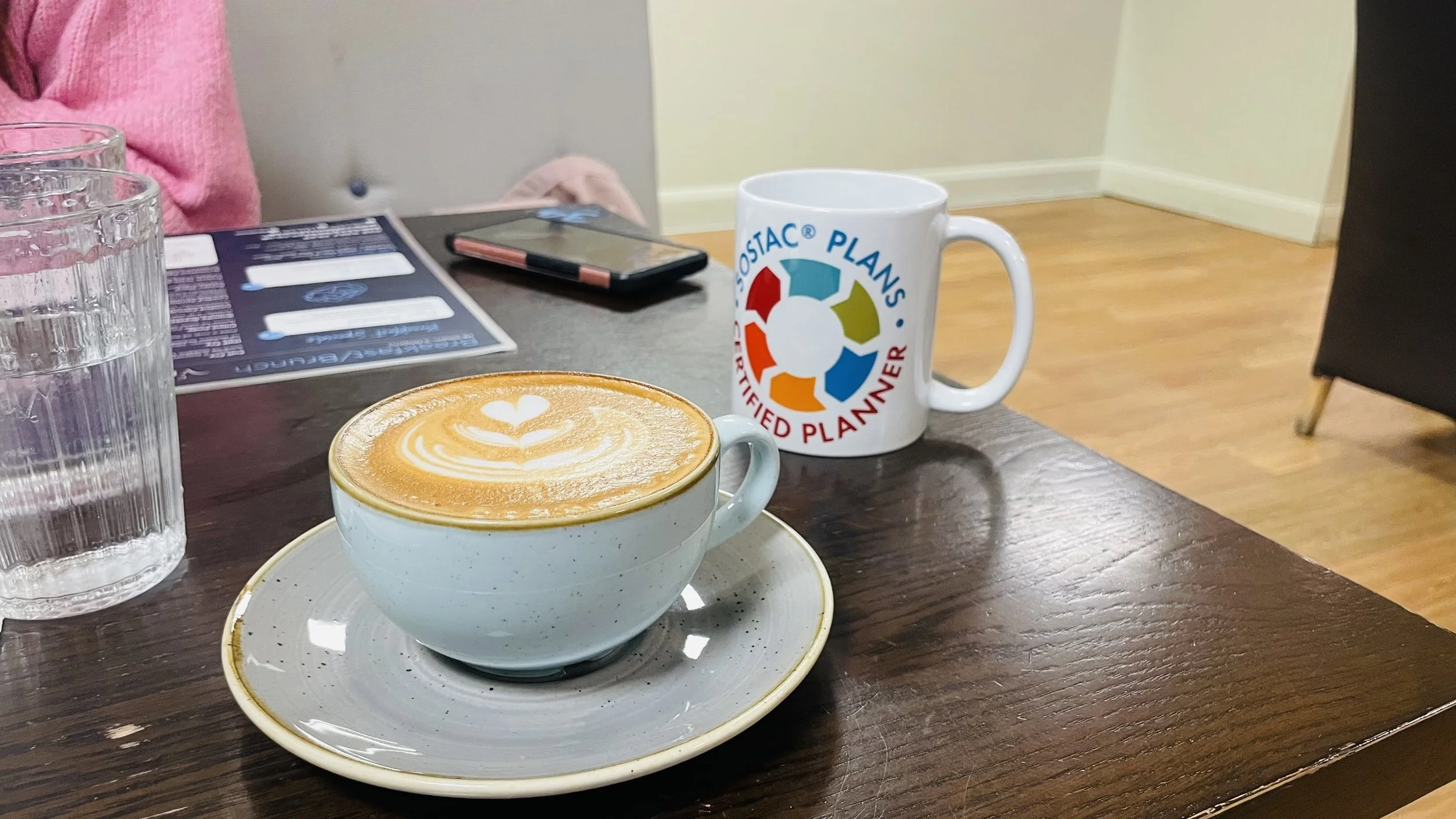 A cup of coffee with latte art, a glass of water, a promotional mug, a smartphone, and a menu on a wooden table in a cafe setting.