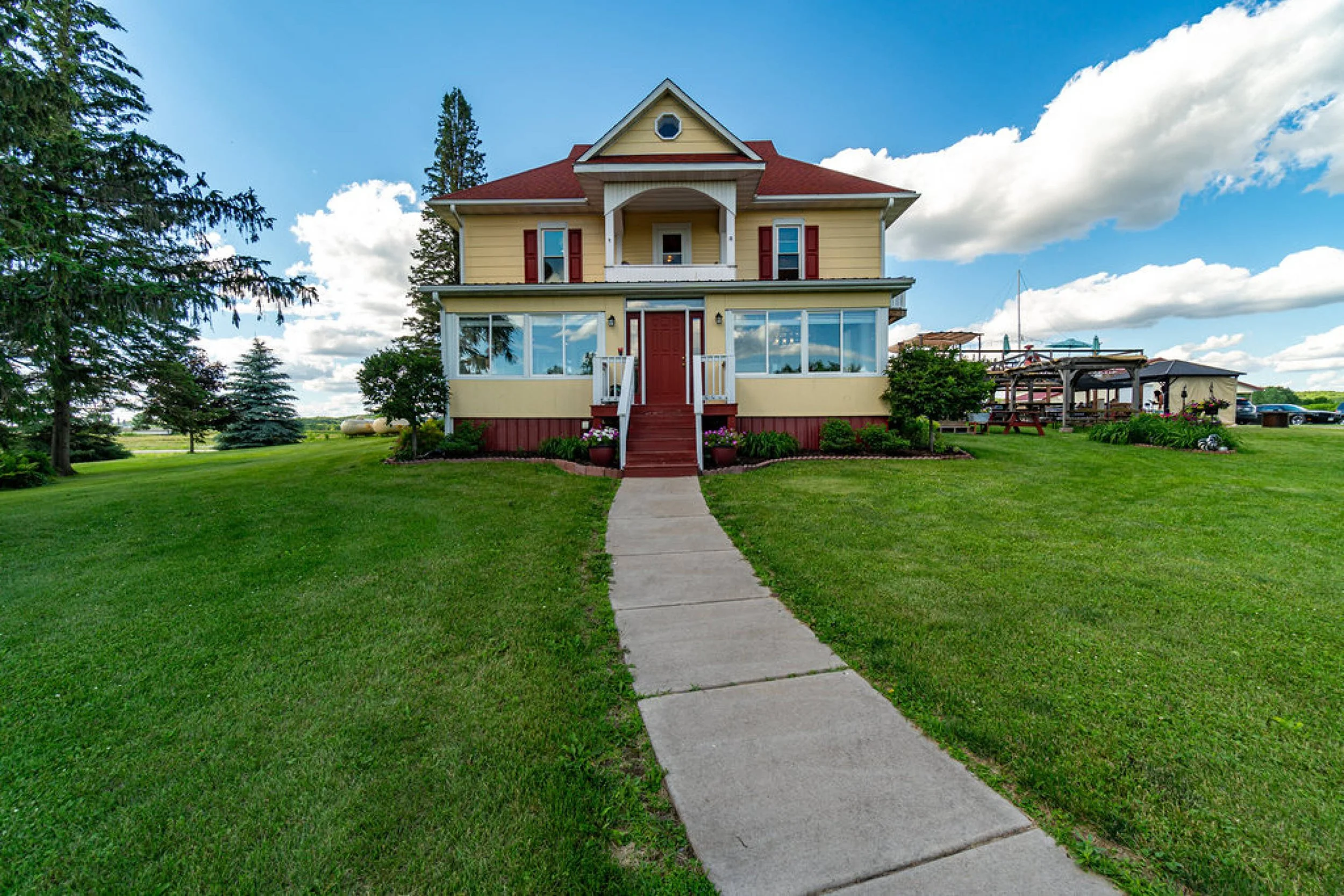 Yellow house with red roof and shutters, white porch, surrounded by green lawn and trees, under a sky with white clouds.