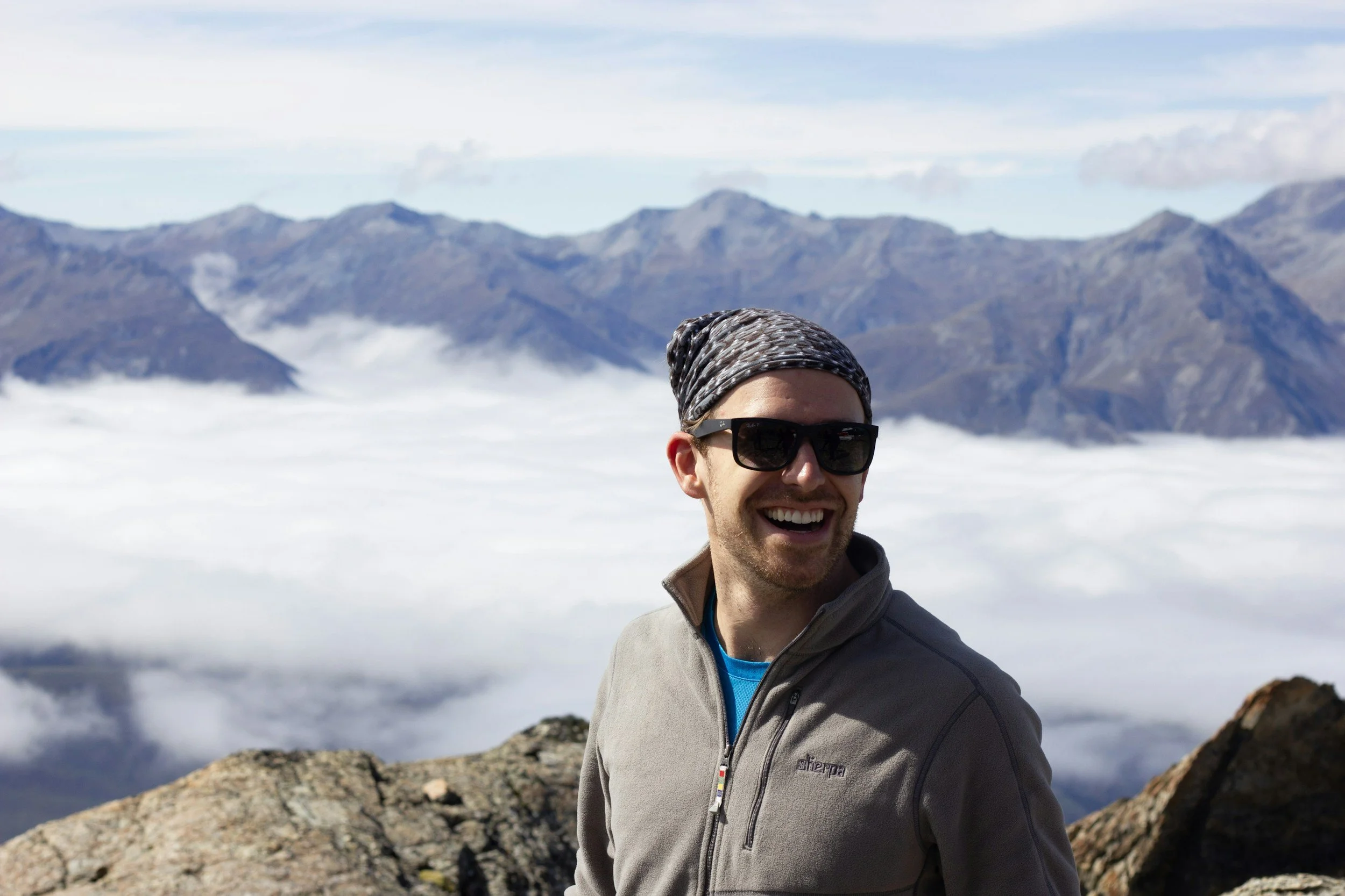 Smiling man wearing sunglasses and a headscarf on a mountain with clouds and distant mountain peaks in the background.