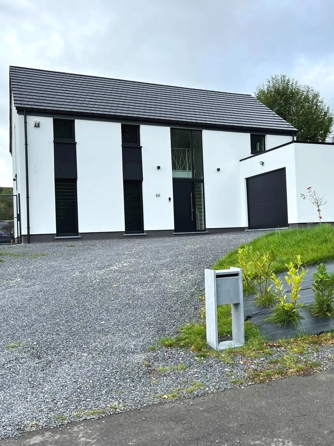 Modern two-story white house with black accents and a dark gray roof, gravel driveway, and small garden with new plants in the front yard.