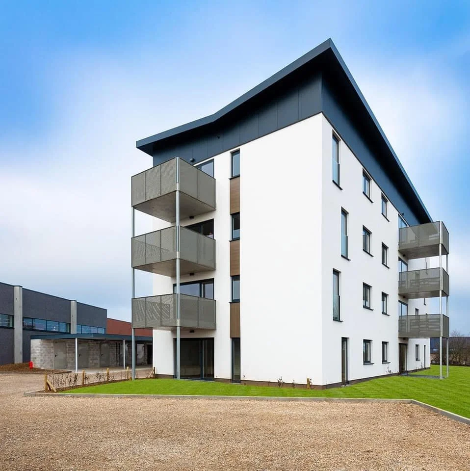 Modern multi-story apartment building with balconies and a sloped roof, surrounded by a grassy area and gravel pathway.