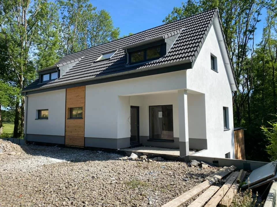 Newly built two-story house with white exterior walls, a dark shingled roof with skylights, and a small front porch, surrounded by trees and construction materials on the ground.