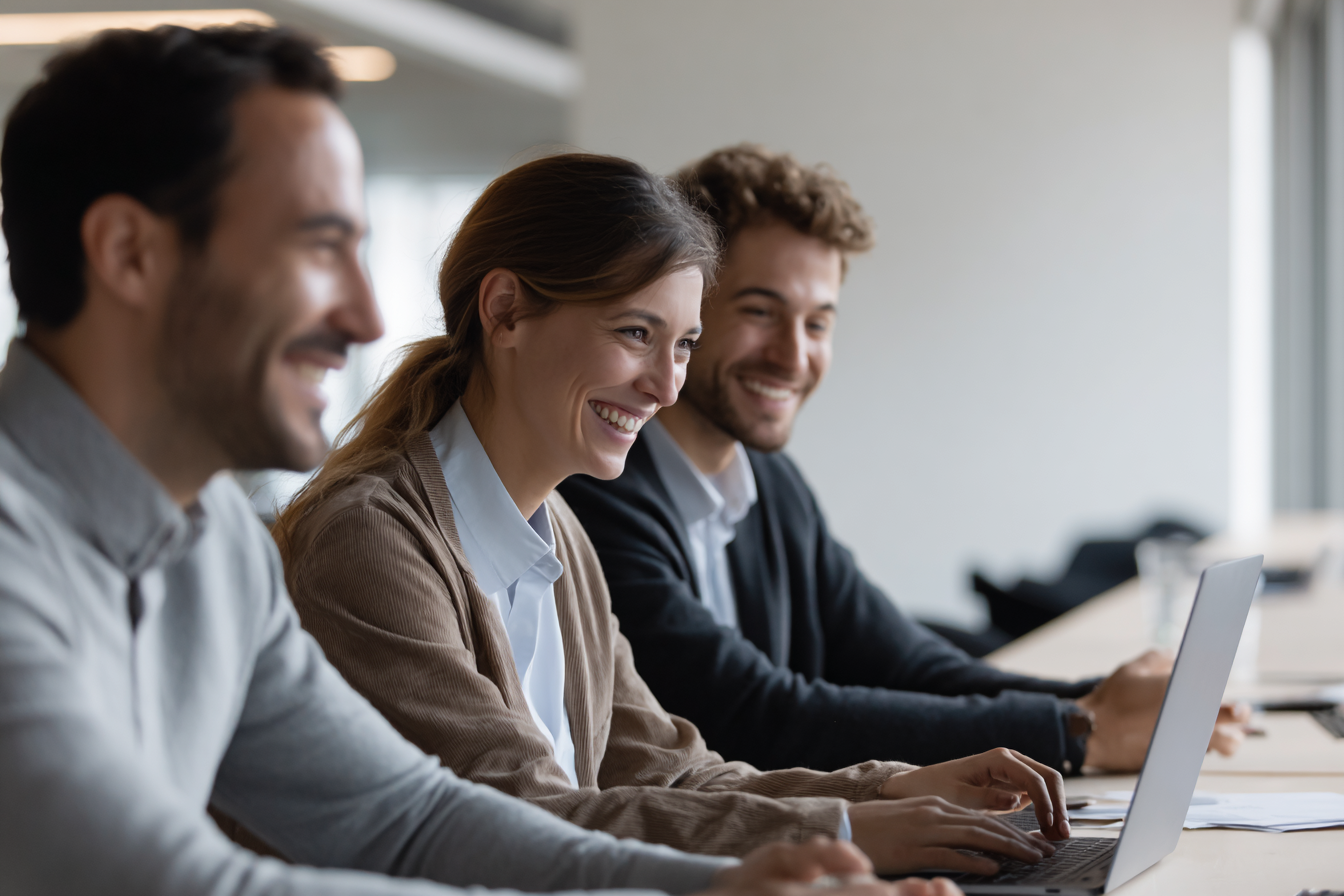 Four diverse people sitting at a conference table, smiling, with laptops in front of them, in a bright, modern office.