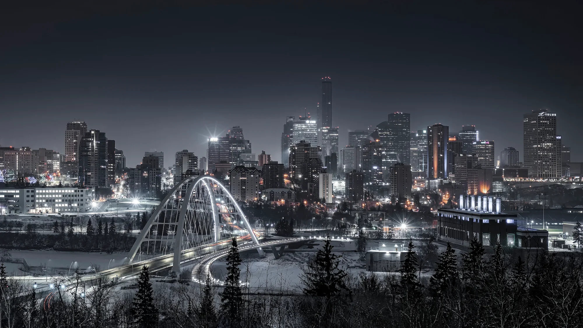 Nighttime city skyline with tall buildings, illuminated bridges, and snow-covered trees in foreground.