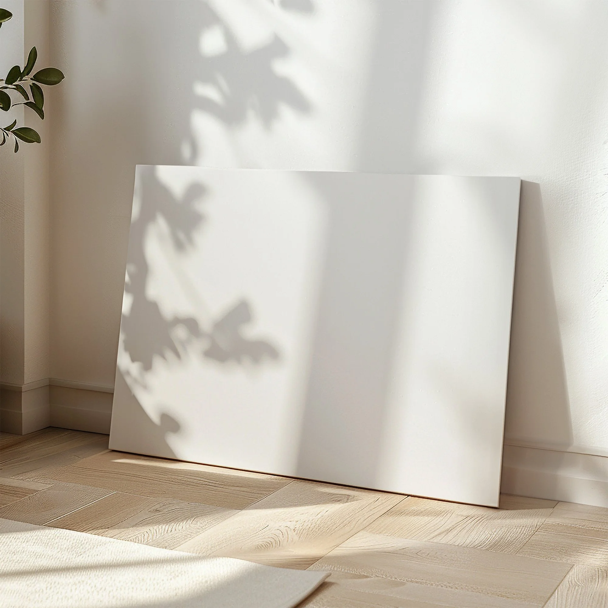 A large blank canvas print leaning against a clean white wall on light oak hardwood floors with soft afternoon tree shadows
