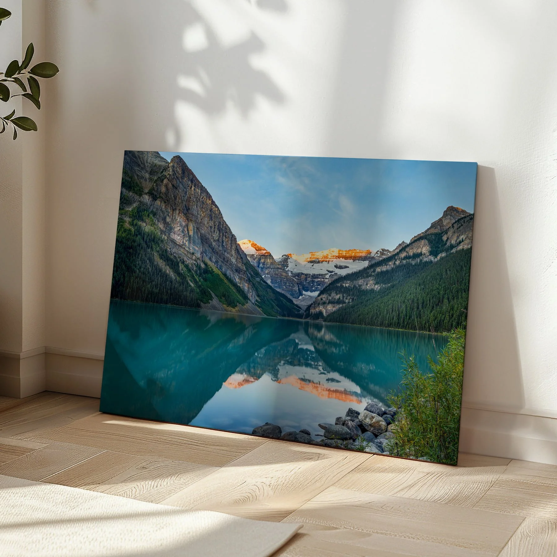 Lake Louise Sunrise gallery-wrapped canvas print leaning in a sunlit room corner on herringbone hardwood flooring, with soft leaf shadows on the wall beside it.