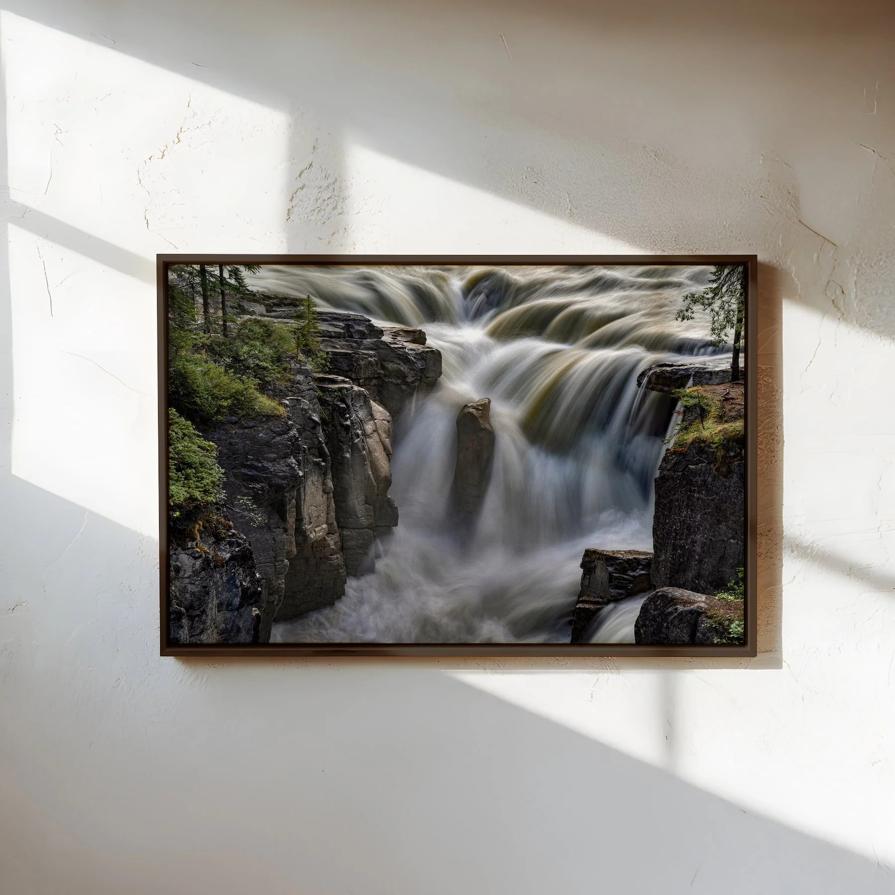 Sunwapta Falls fine art photograph in a walnut floating canvas frame, displayed on a textured white wall with natural window light and shadows.