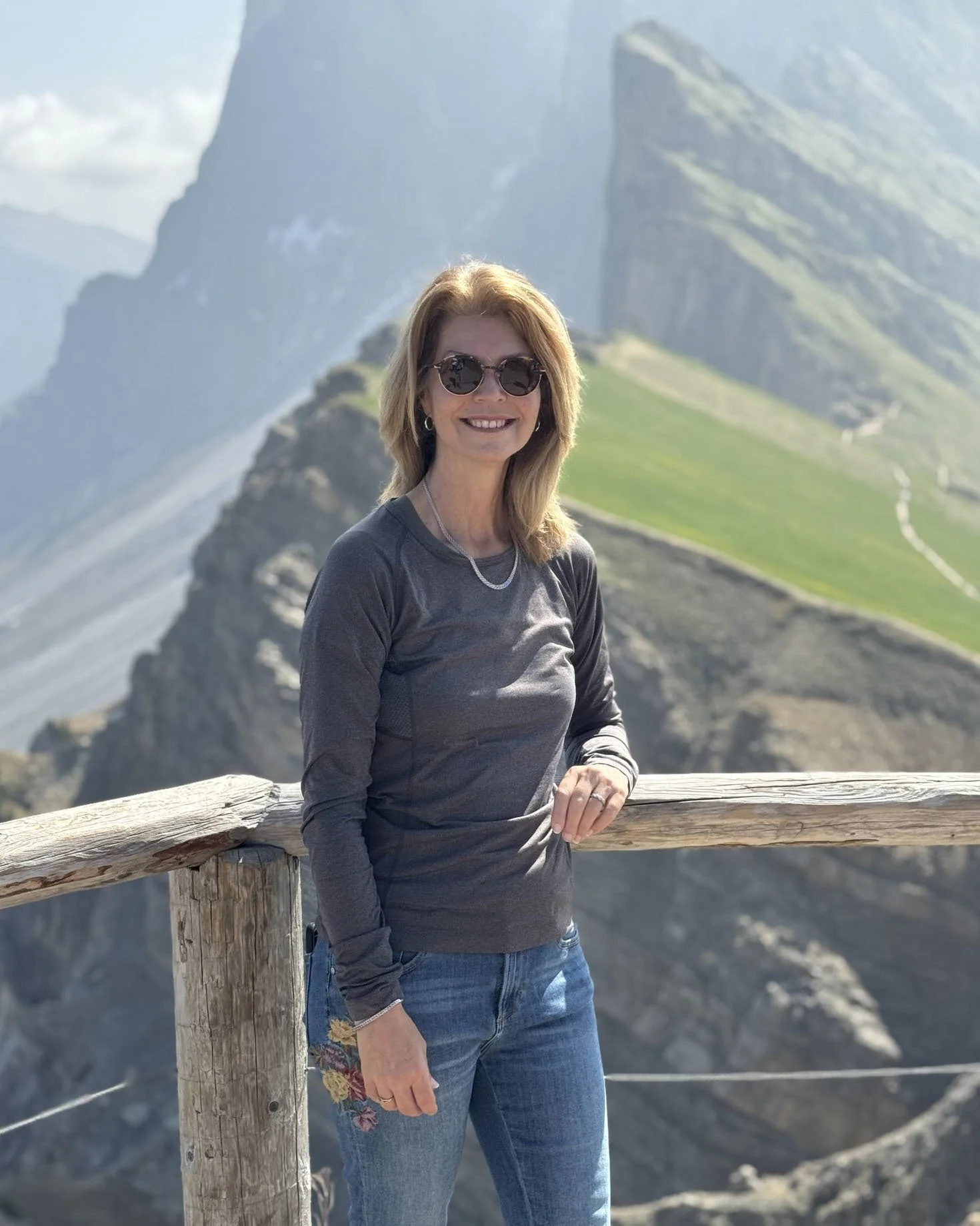 Lee-Ann with sunglasses smiling, standing by a wooden railing in front of Seceda, a mountainous landscape with rocky peaks and green grassy slopes.