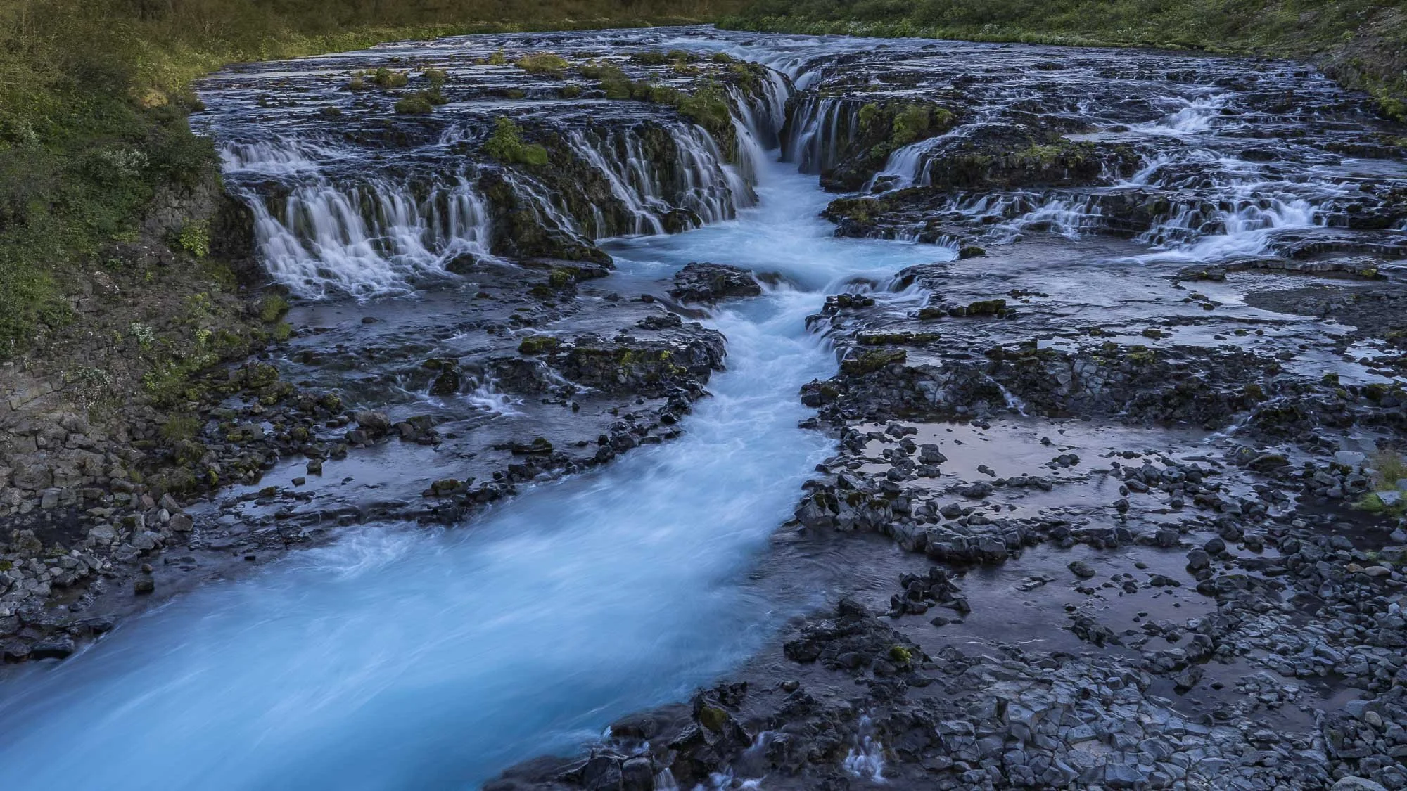 Turquoise water flowing over dark volcanic rock and converging through the centre of Brúarfoss waterfall in Iceland.