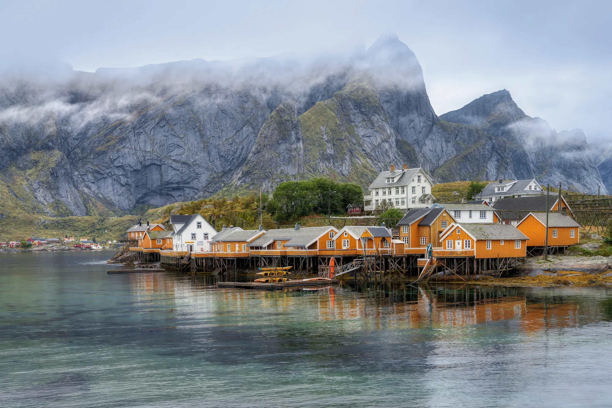 A scenic view of colorful houses on stilts by the water, with steep, rocky mountains in the background, under a cloudy sky.