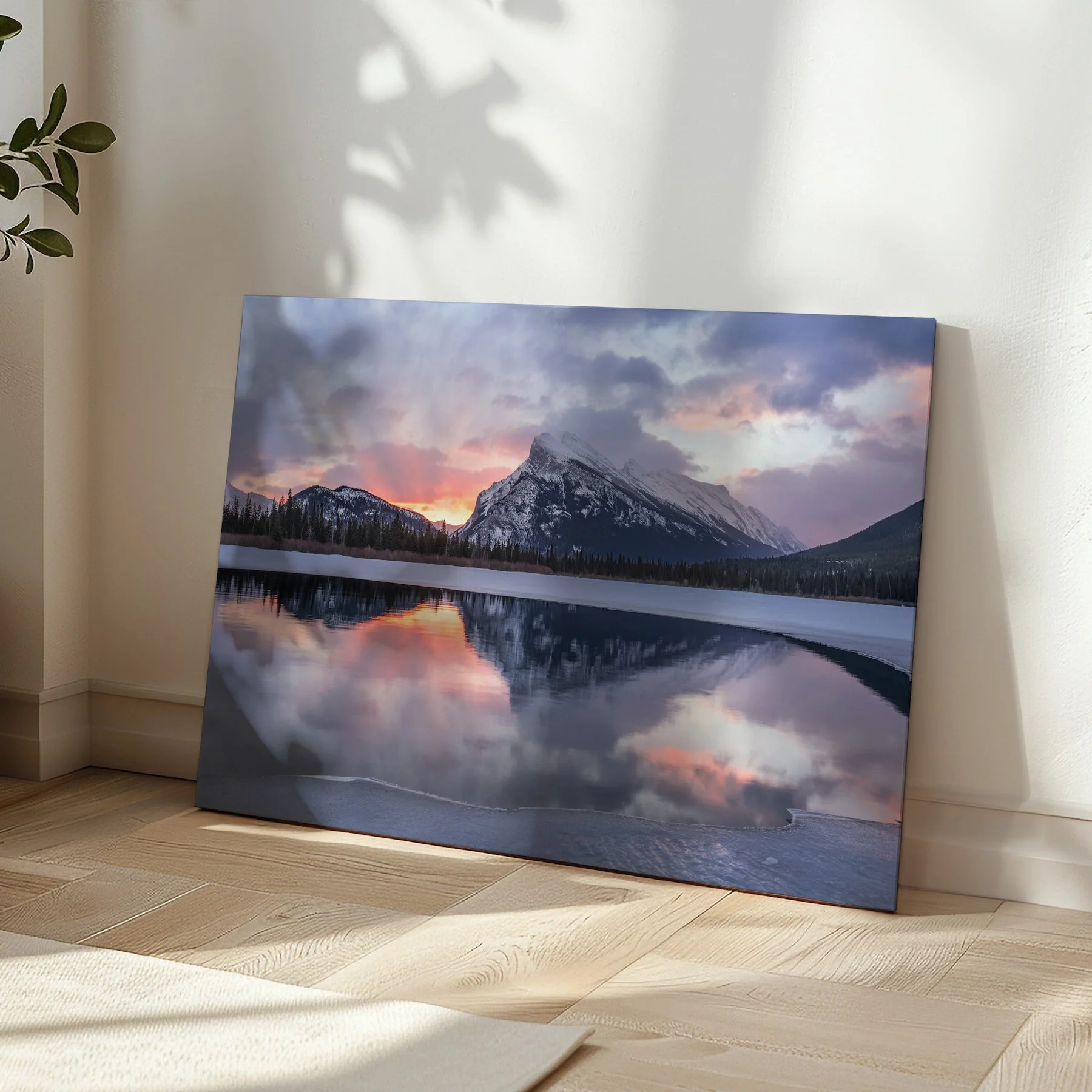 Mount Rundle Reflections gallery-wrapped canvas print leaning in a sunlit room corner on herringbone hardwood flooring, with soft leaf shadows on the wall beside it.