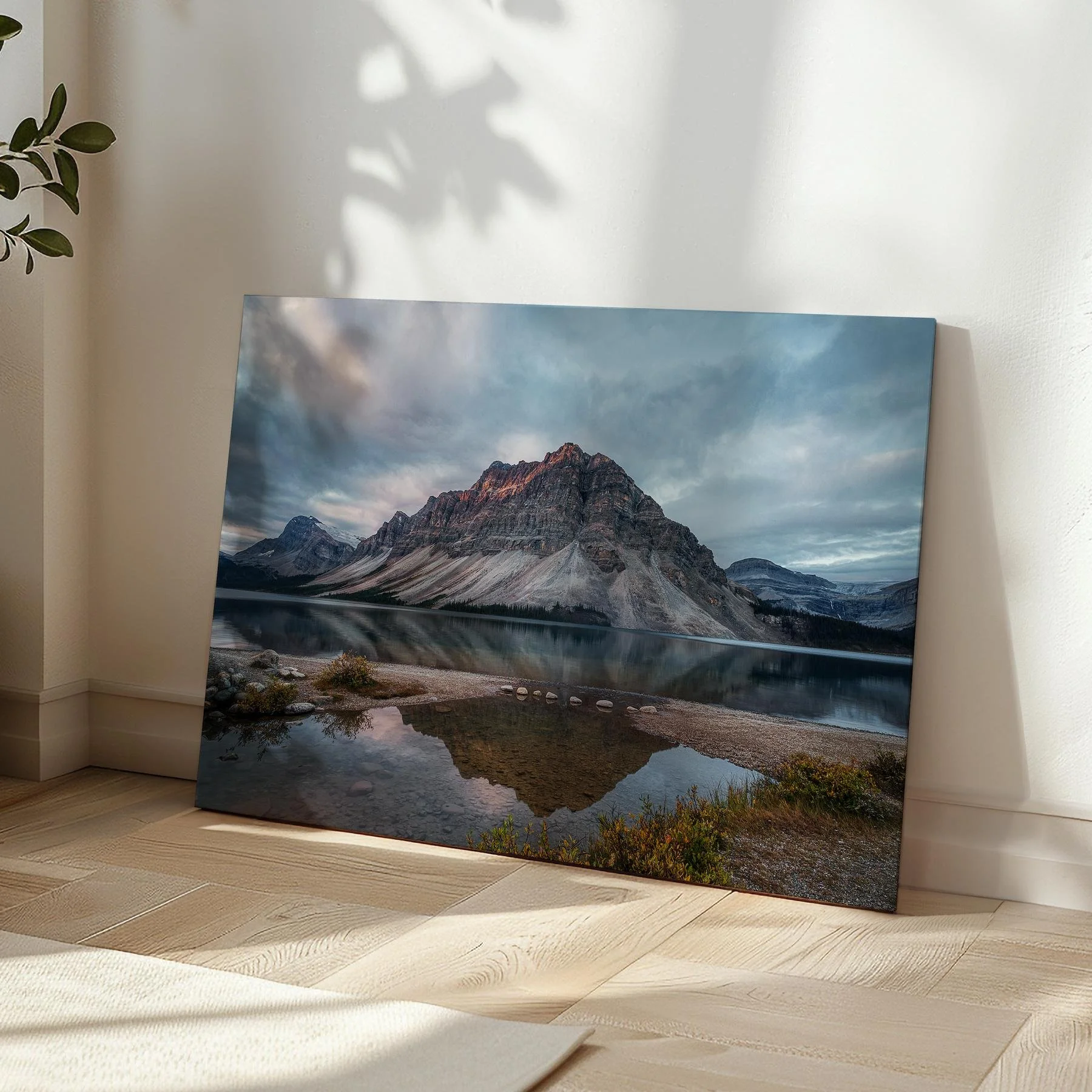 Bow Lake Reflections gallery-wrapped canvas print leaning in a sunlit room corner on herringbone hardwood flooring, with soft leaf shadows on the wall beside it.
